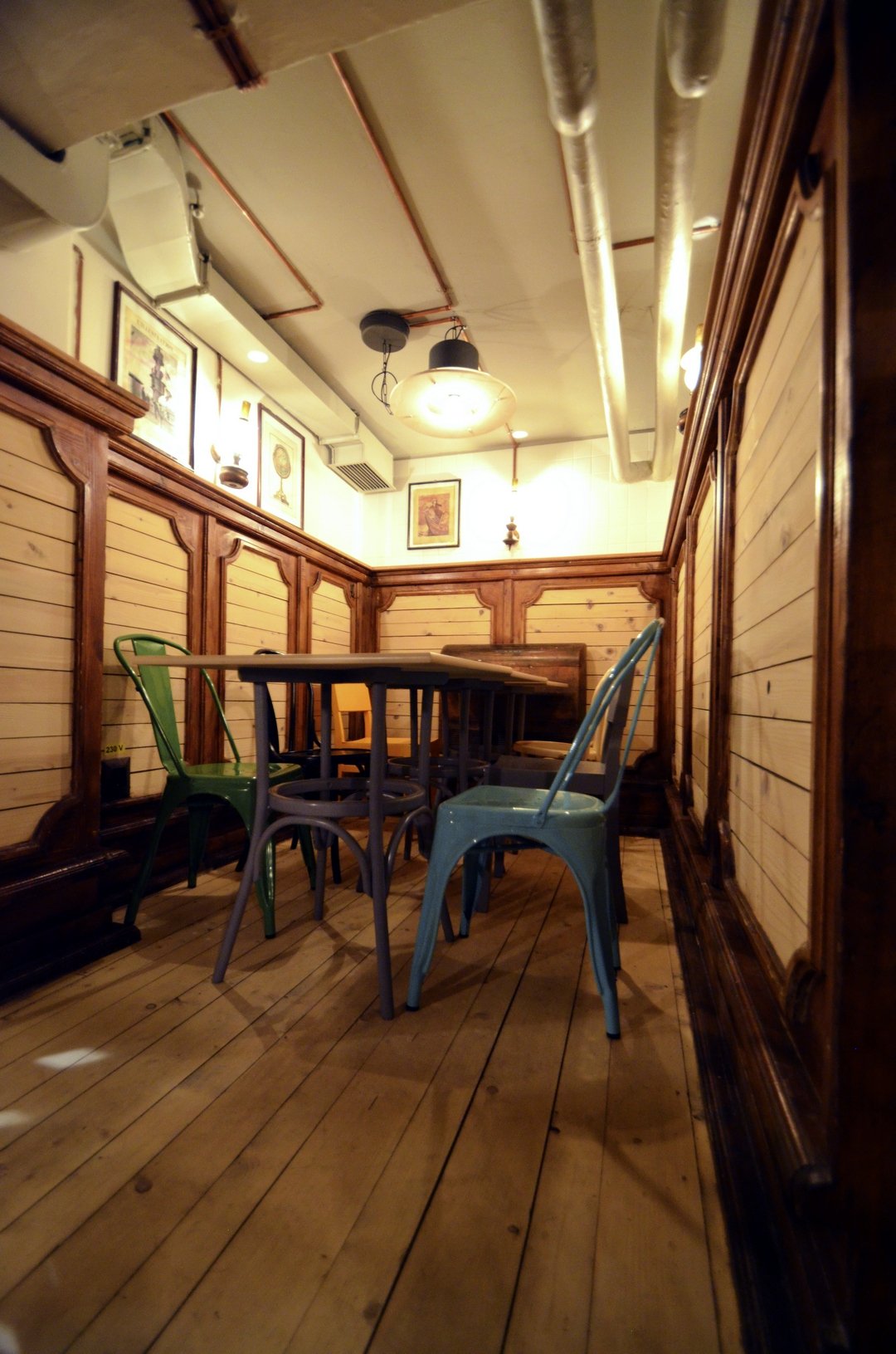 Cozy cafe corner with a wooden floor, wooden paneled walls, and colorful metal chairs around a small table. Framed pictures hang on the wall, and warm lighting illuminates the space.