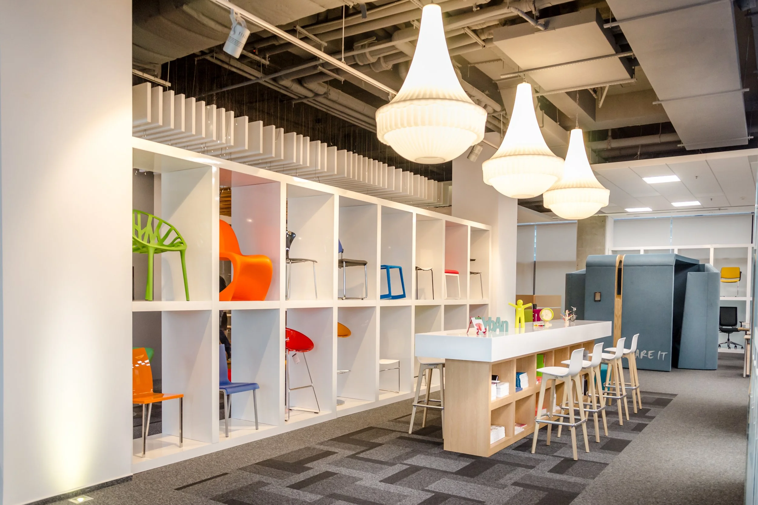 Modern office or retail space with a display of colorful chairs arranged in a white shelving unit, three large white pendant lights hanging from the ceiling, a long white table with white chairs, and a blue booth-like structure in the background.