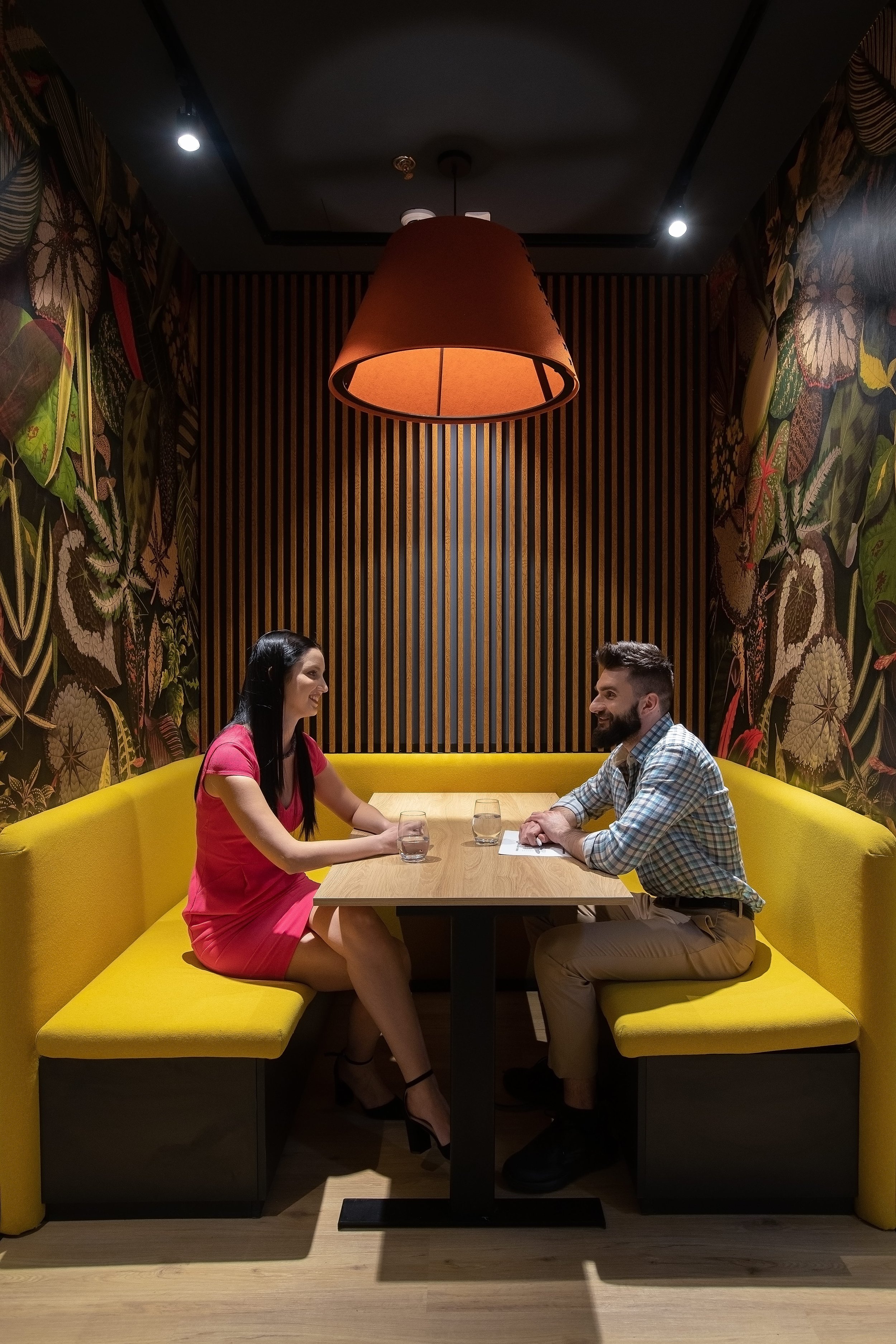 A woman and a man sit across from each other in a restaurant booth with yellow seating, engaged in conversation. A large pendant lamp hangs above them, and the walls have floral and botanical wallpaper.