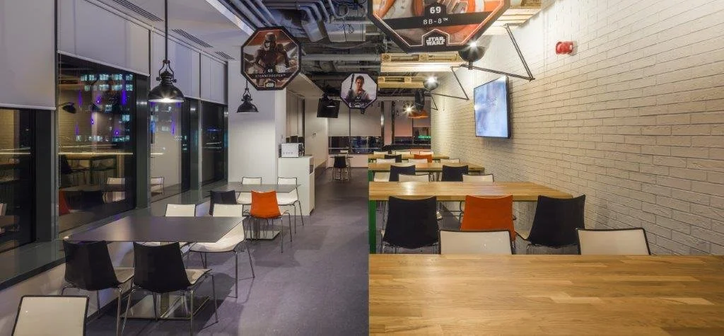 Empty modern cafe or restaurant with a combination of black, white, and wooden tables and chairs; large glass windows on the left; TV screen on the right wall; exposed ceiling pipes; and decorative wall art with popular sci-fi characters.