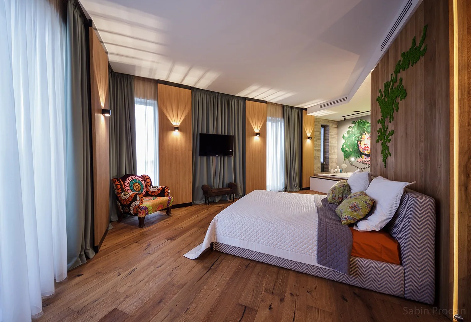 Modern bedroom with wooden flooring, large bed with patterned headboard and decorative pillows, colorful armchair, wall-mounted TV, large windows with curtains, and a mural of a woman's face surrounded by greenery.
