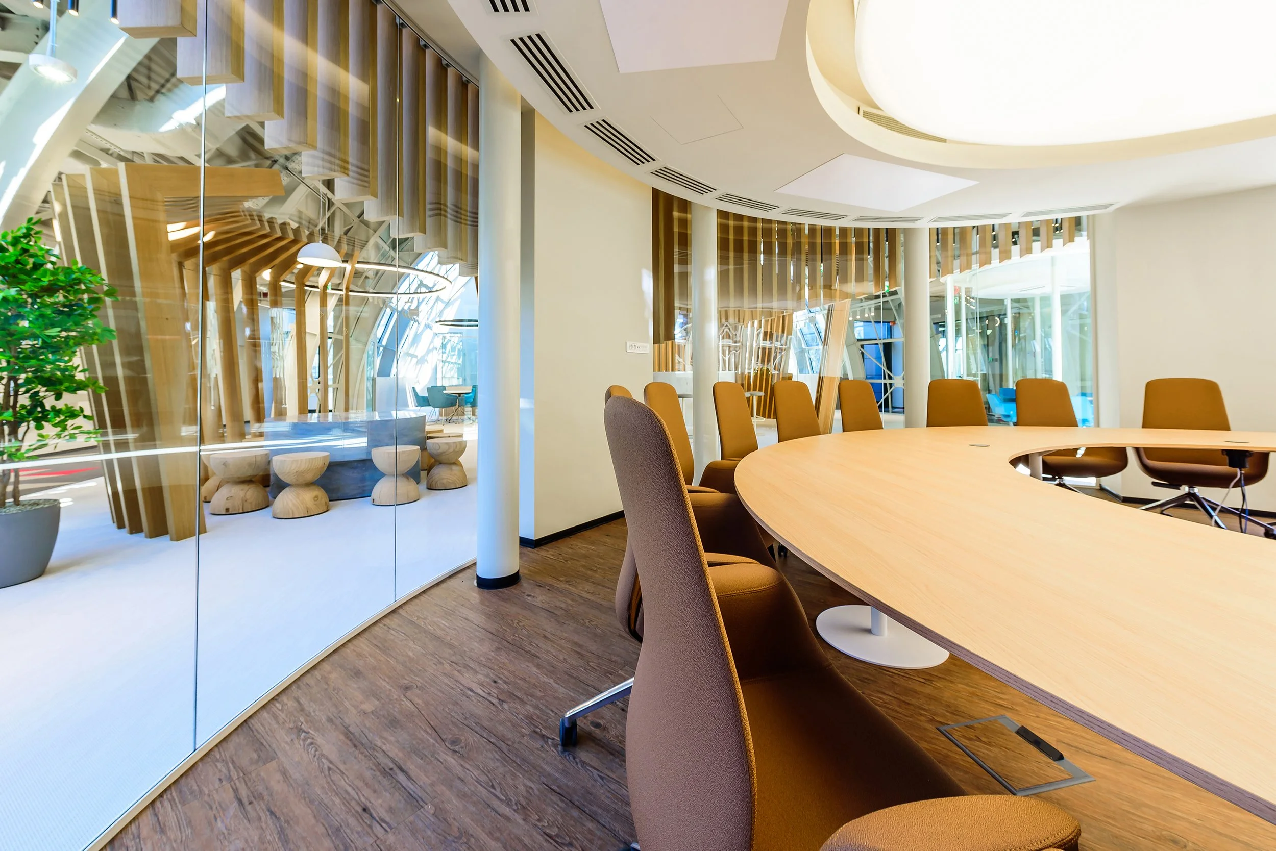 Modern conference room with a large curved wooden table and brown chairs, glass walls with a view of a bright, contemporary outdoor seating area with wooden benches, green plants, and architectural wooden structures.