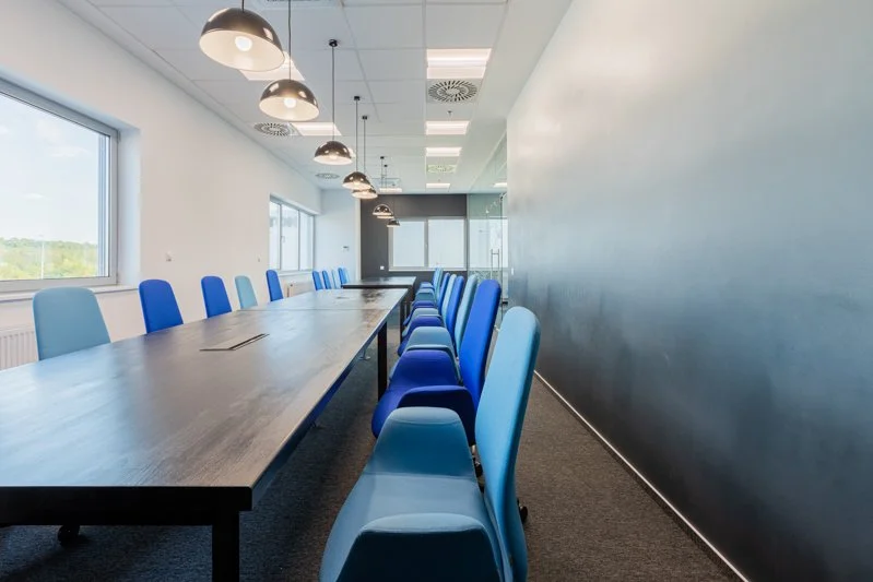Empty conference room with a long table and alternating light blue and dark blue chairs, large windows on one side, and hanging ceiling lights.