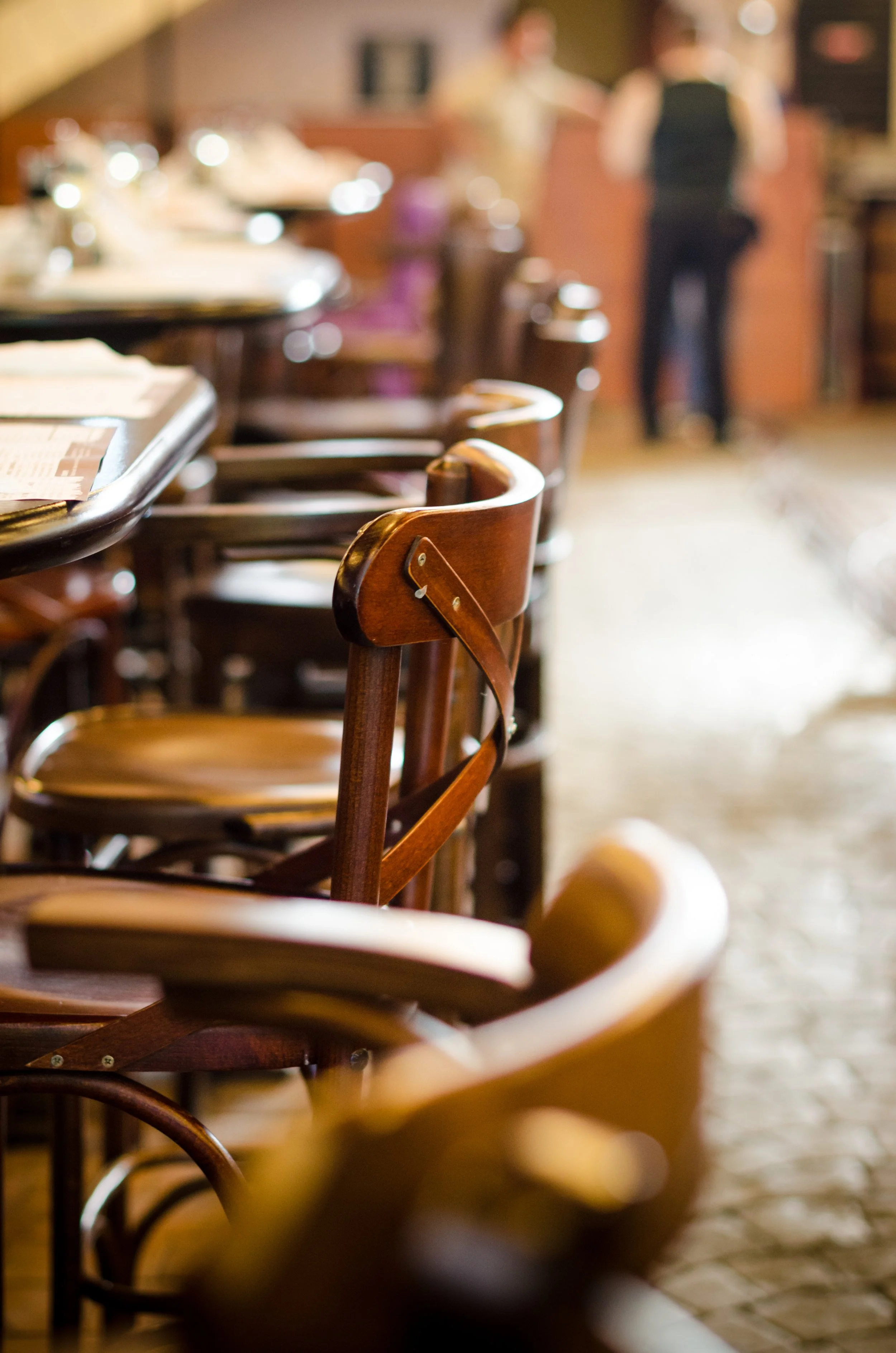 Rows of empty wooden chairs and tables in a restaurant, with blurred people in the background.