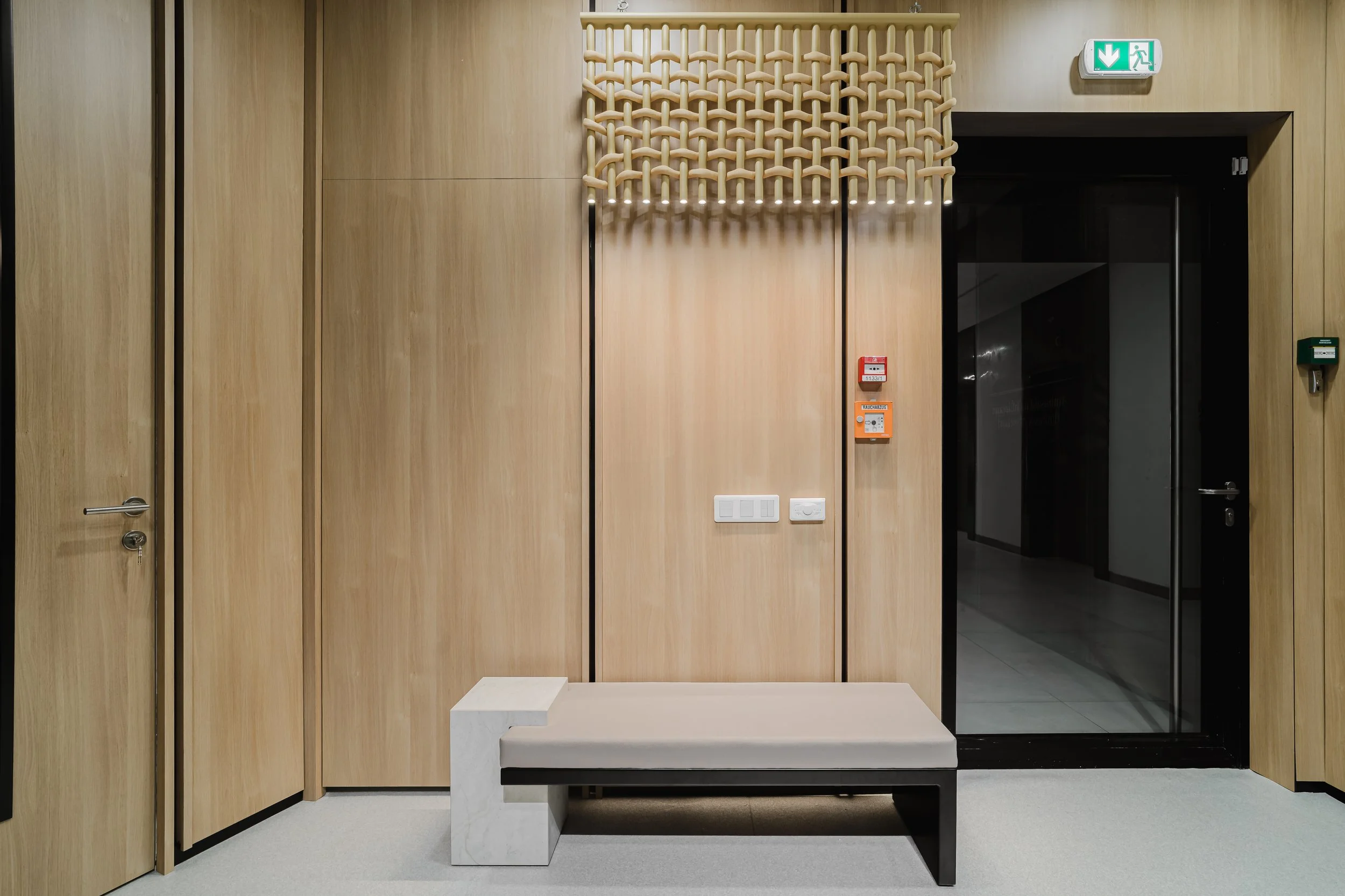 Modern waiting area with light wood-paneled walls, a beige bench with black legs, a white marble side table, a decorative wooden lattice piece, and an emergency exit sign above a black door.