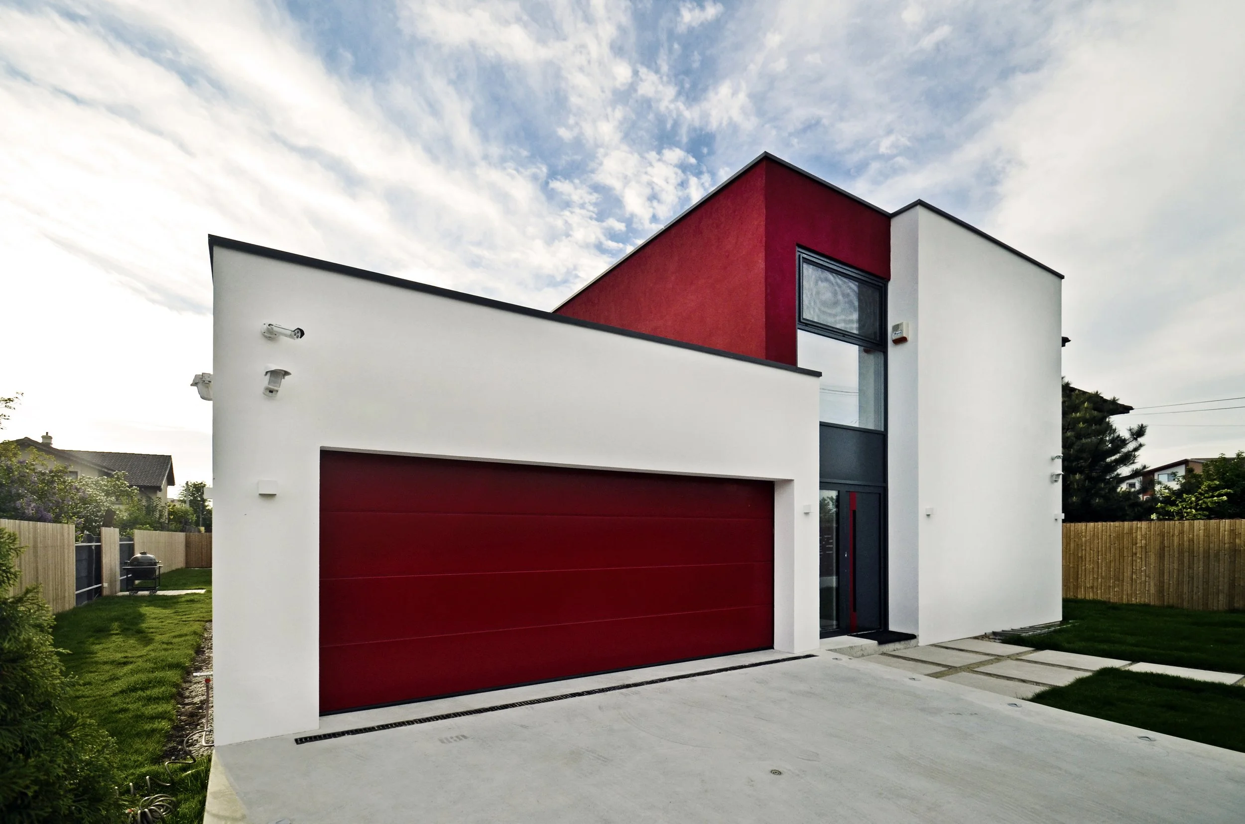 Modern two-story house with a white and red exterior, large garage door, and a front yard with a concrete driveway and green lawn, under a partly cloudy sky.
