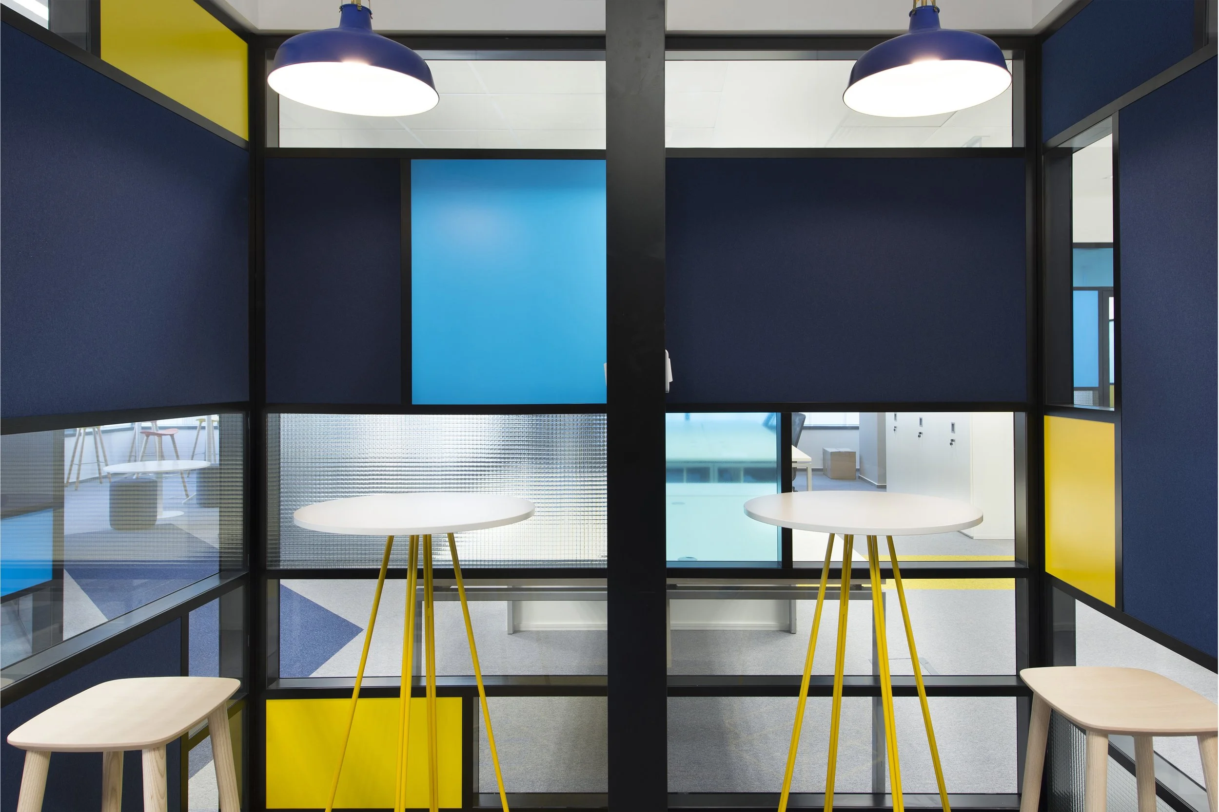 Modern office partitions with blue and yellow panels, glass windows, white tables, and wooden stools, with ceiling lights overhead.