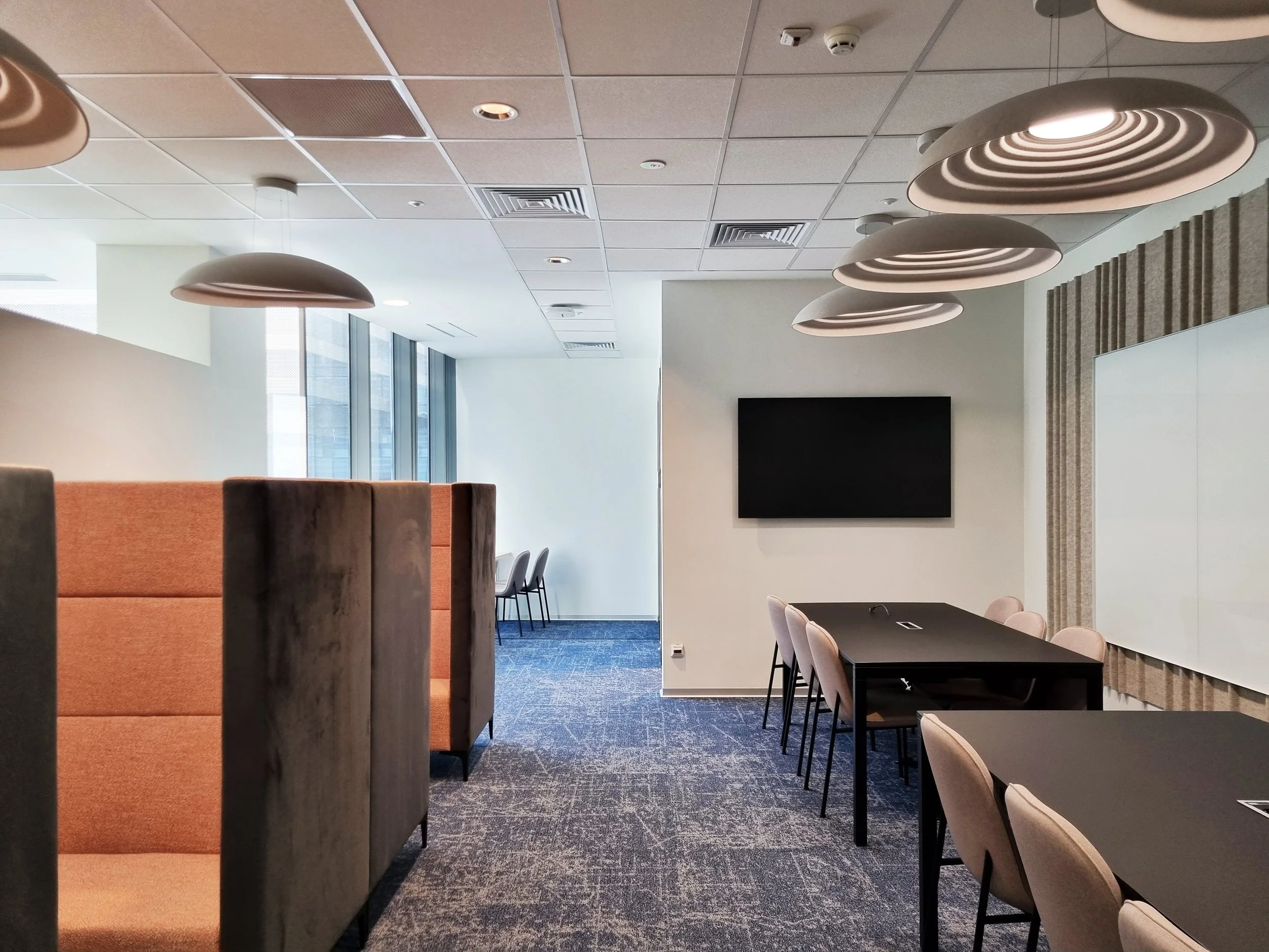 Empty modern conference room with black tables, beige chairs, a large whiteboard, and a wall-mounted television, illuminated by natural light from tall windows and ceiling lights.