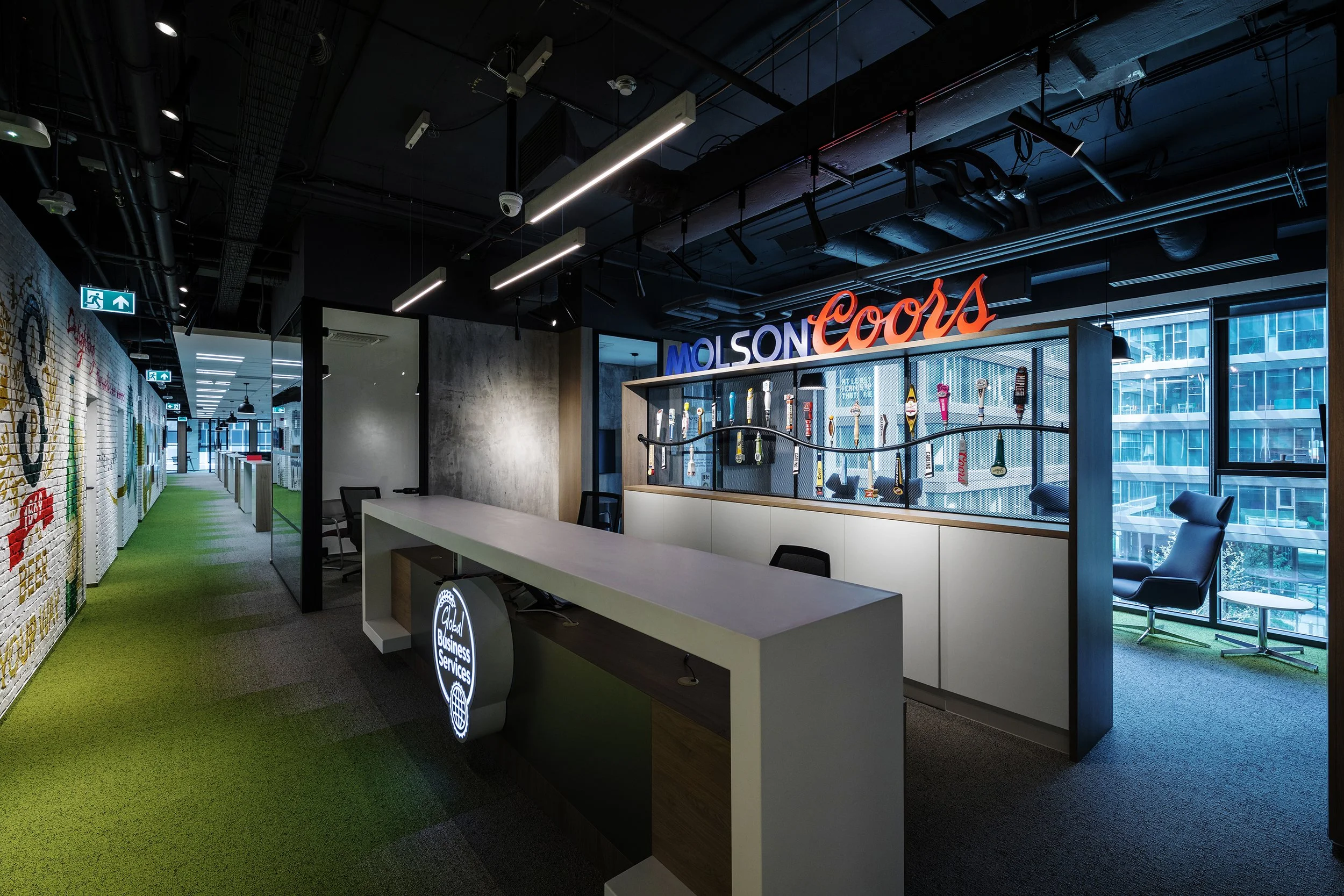Modern office reception area with a white desk, black chairs, a display of beer taps, and a neon sign reading 'Molson Coors' against large windows that show city buildings outside.