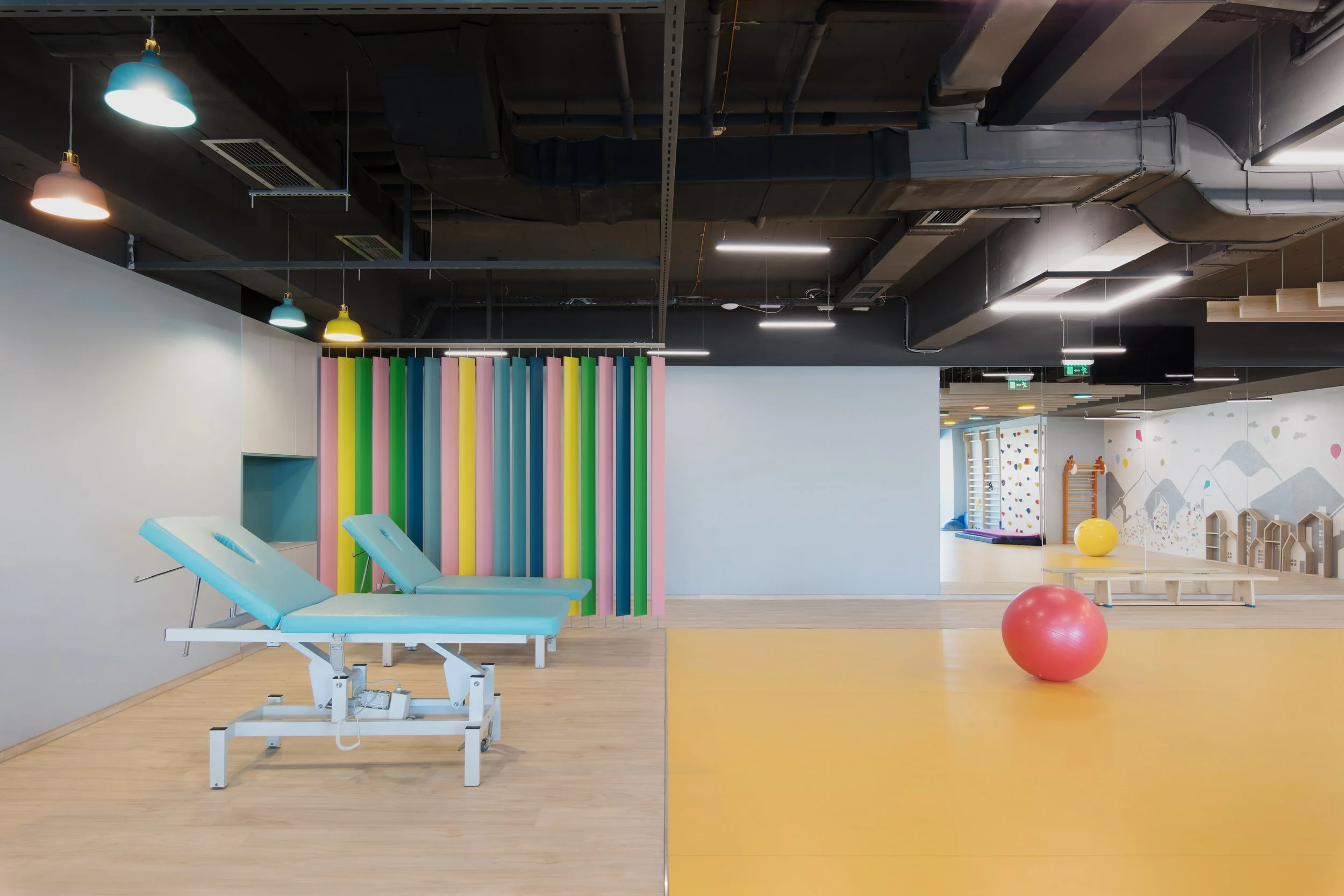 A brightly lit indoor gym or physical therapy space with two examination tables, one on each side of a colorful vertical striped wall. To the right, there is a yellow exercise ball, a pink exercise ball, and other play or therapy equipment in a room with a mountain and house mural on the wall.