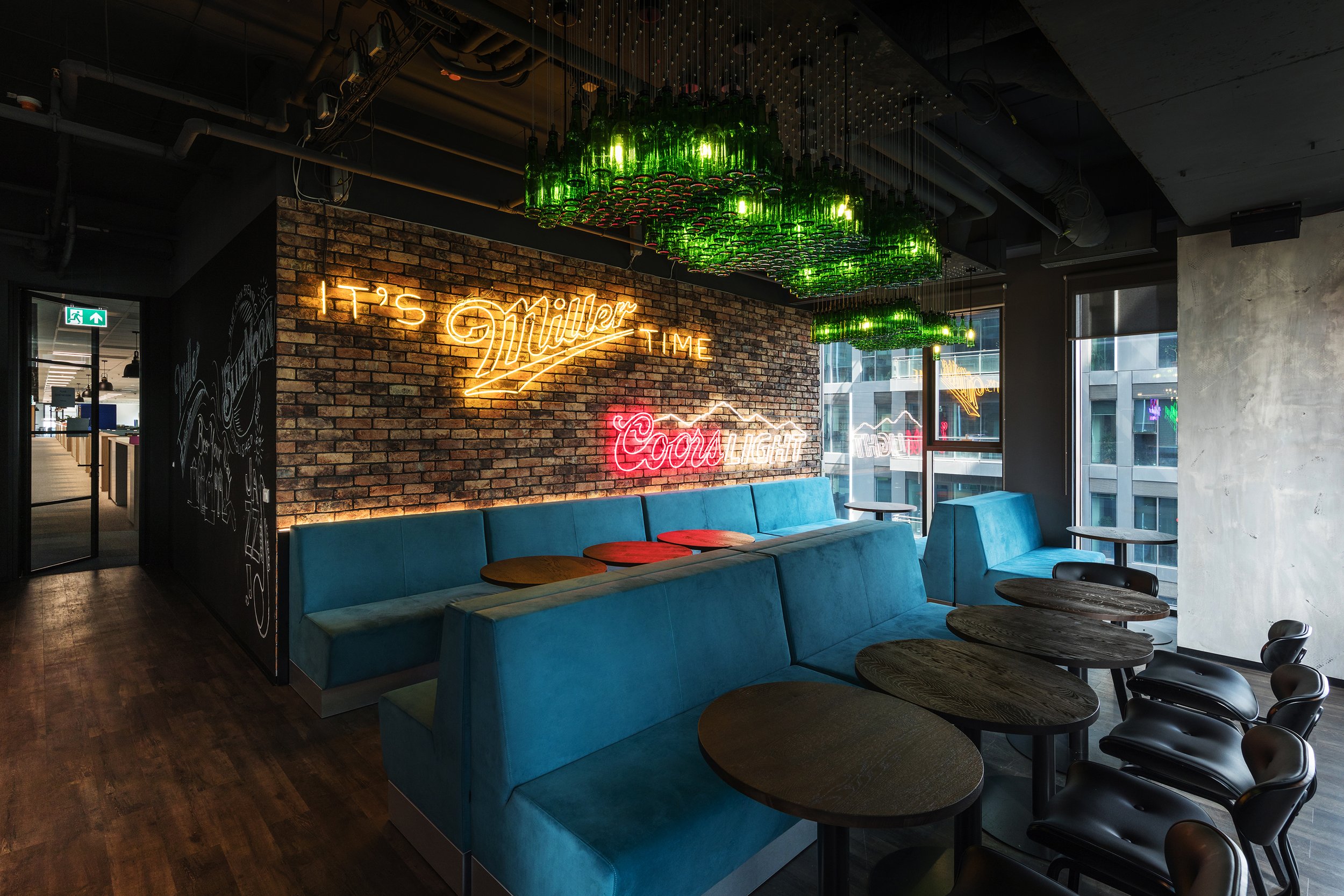 Modern bar or restaurant interior with brick wall, blue seating, and green glass bottle chandelier, neon signs reading 'It's Miller Time' and 'Coca Cola'.