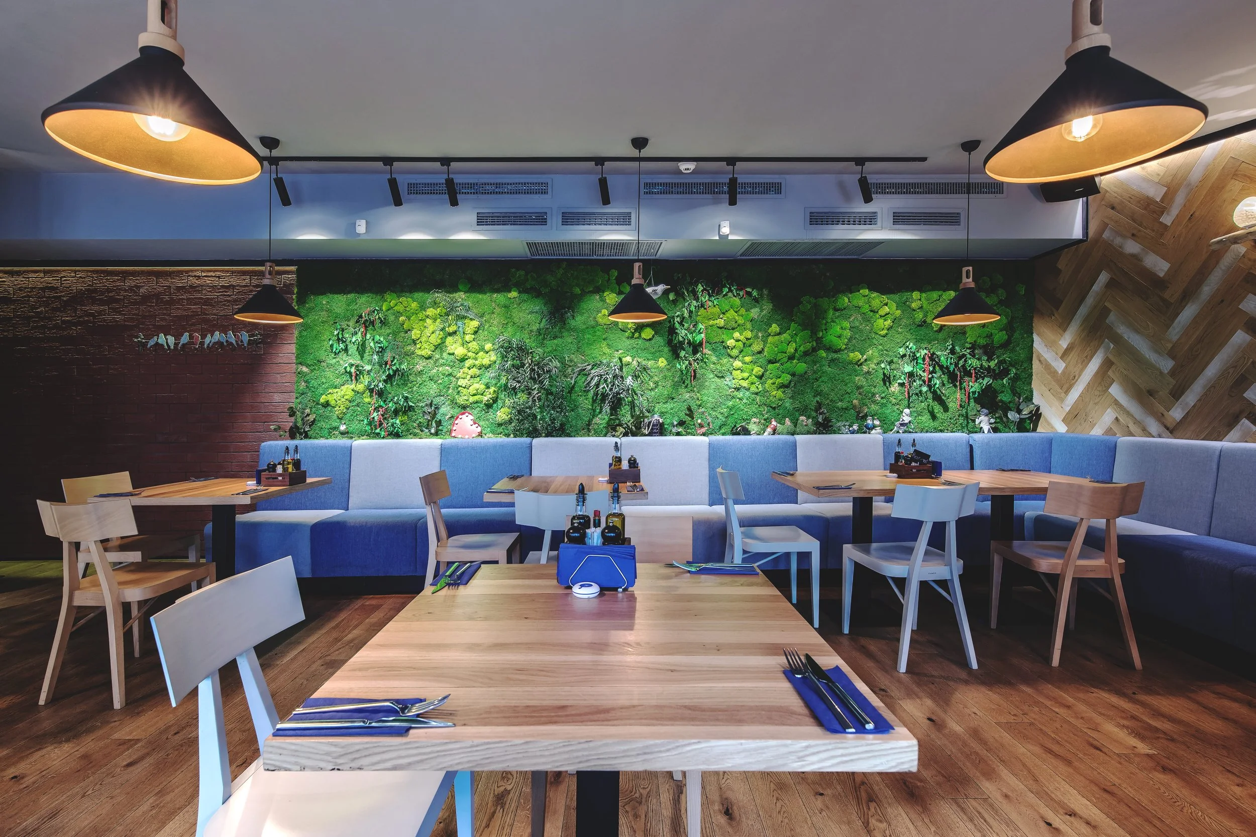 Empty restaurant with wooden tables, white and light wood chairs, a green wall with plants, hanging black pendant lights, and wooden wall accents.