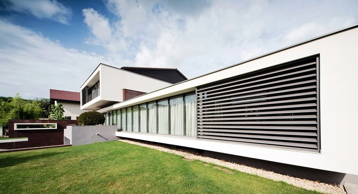 Modern two-story house with large windows, white exterior, and contemporary design, set on a green lawn under a partly cloudy sky.