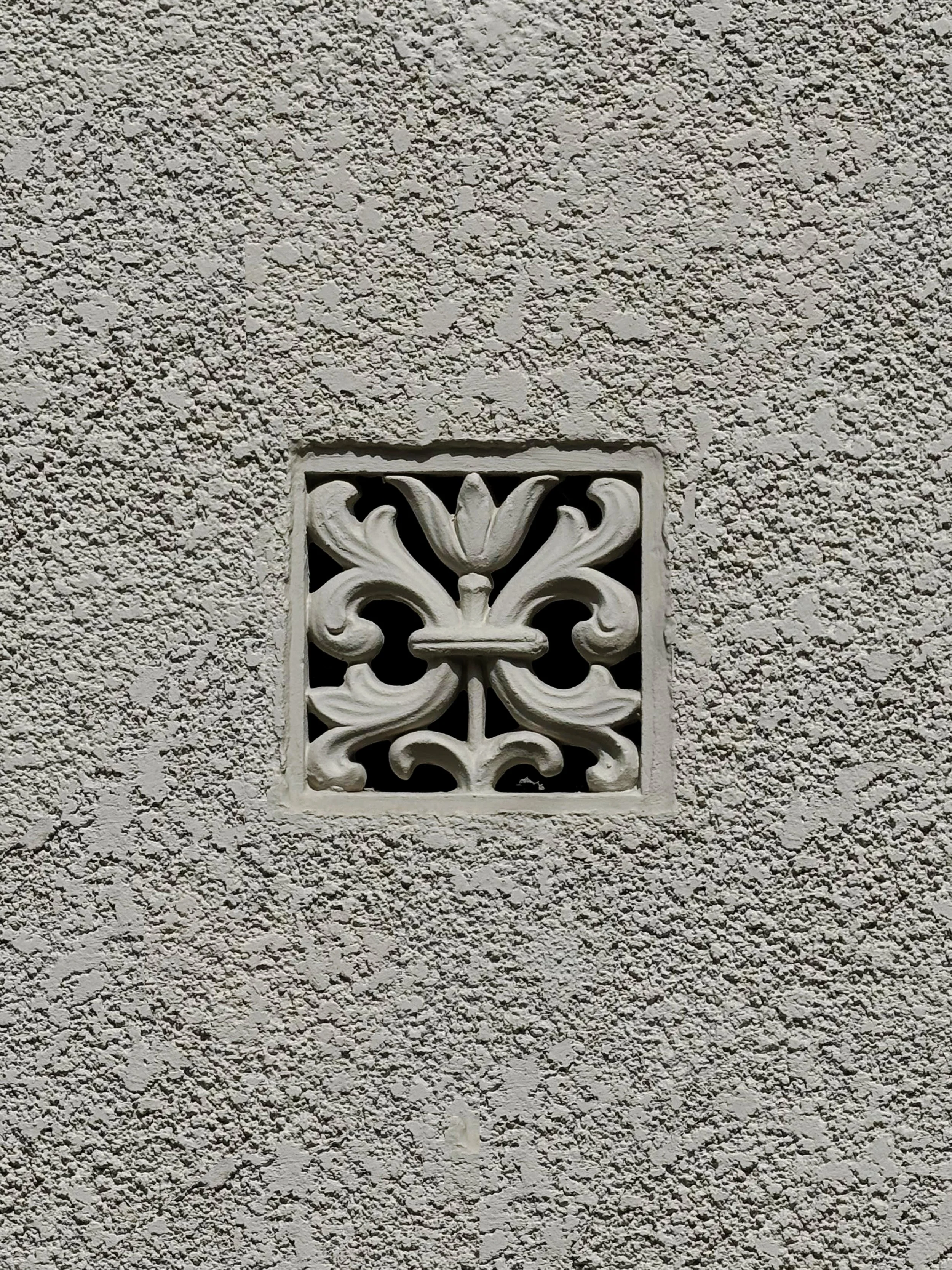 A decorative white metal vent cover with an ornate floral design, mounted on a textured white stucco wall.