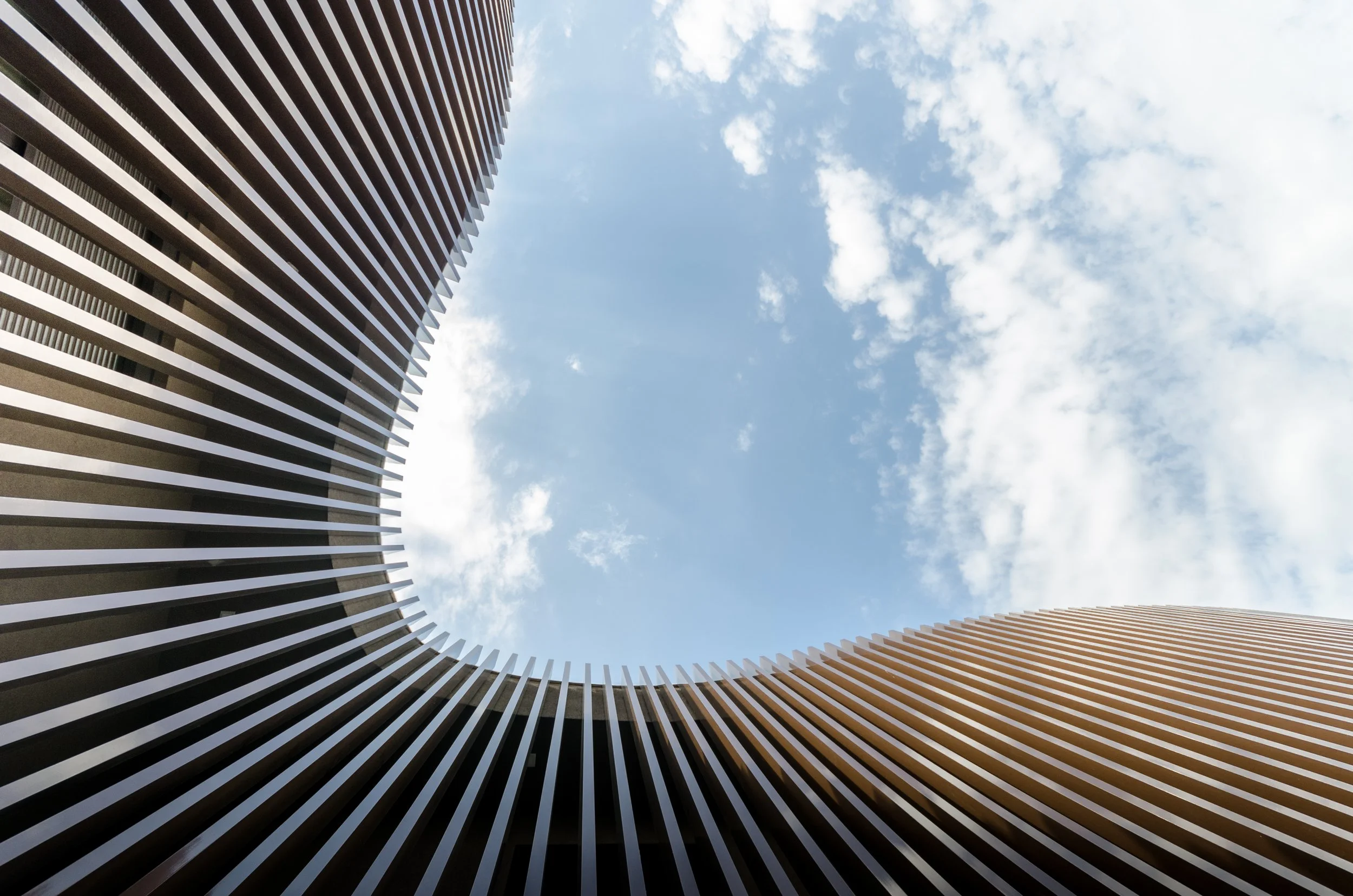 Looking up at modern architectural building with vertical wooden slats curved around open sky with clouds.