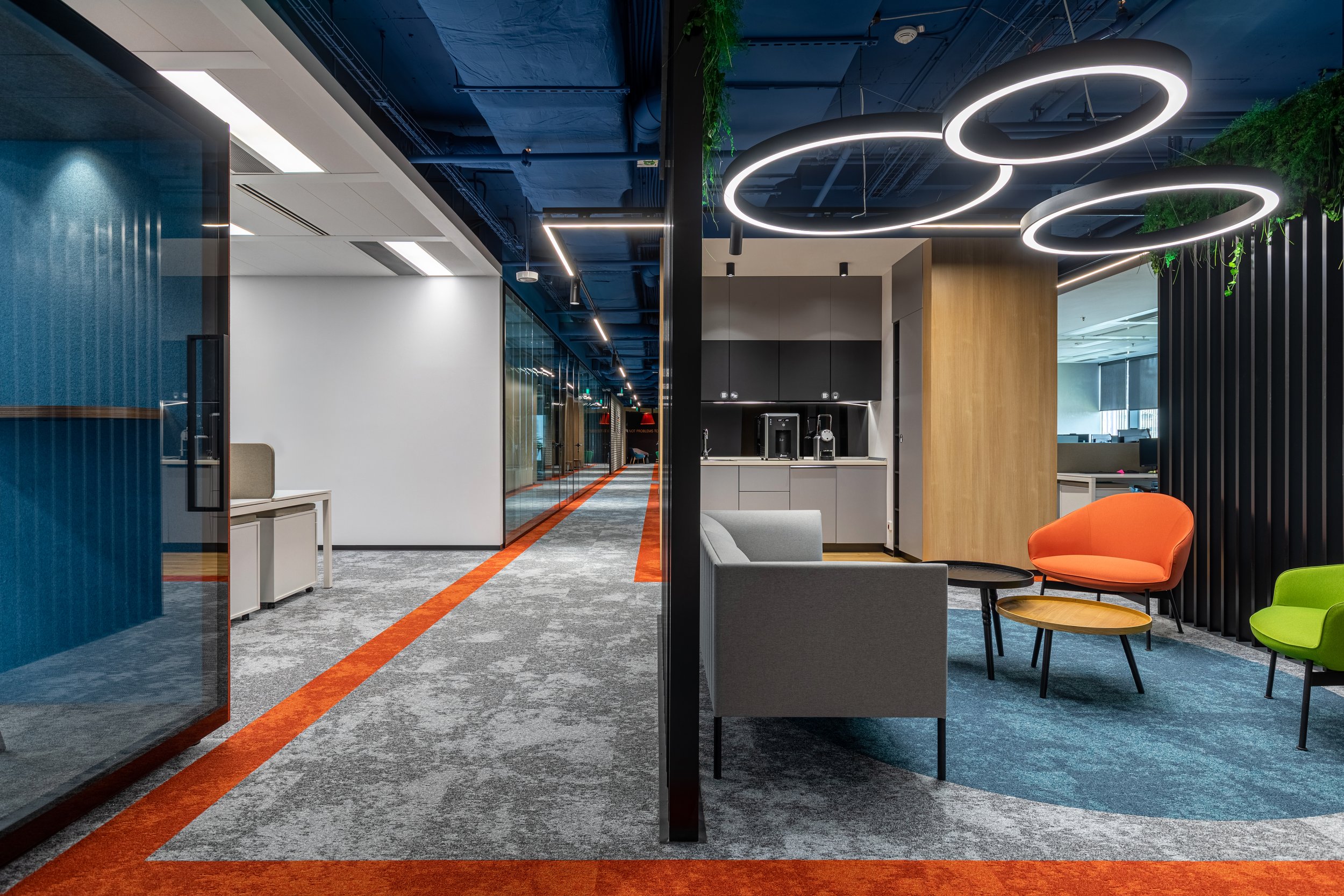 Modern office lounge with colorful chairs, circular ceiling lights, a coffee station, and glass walls.