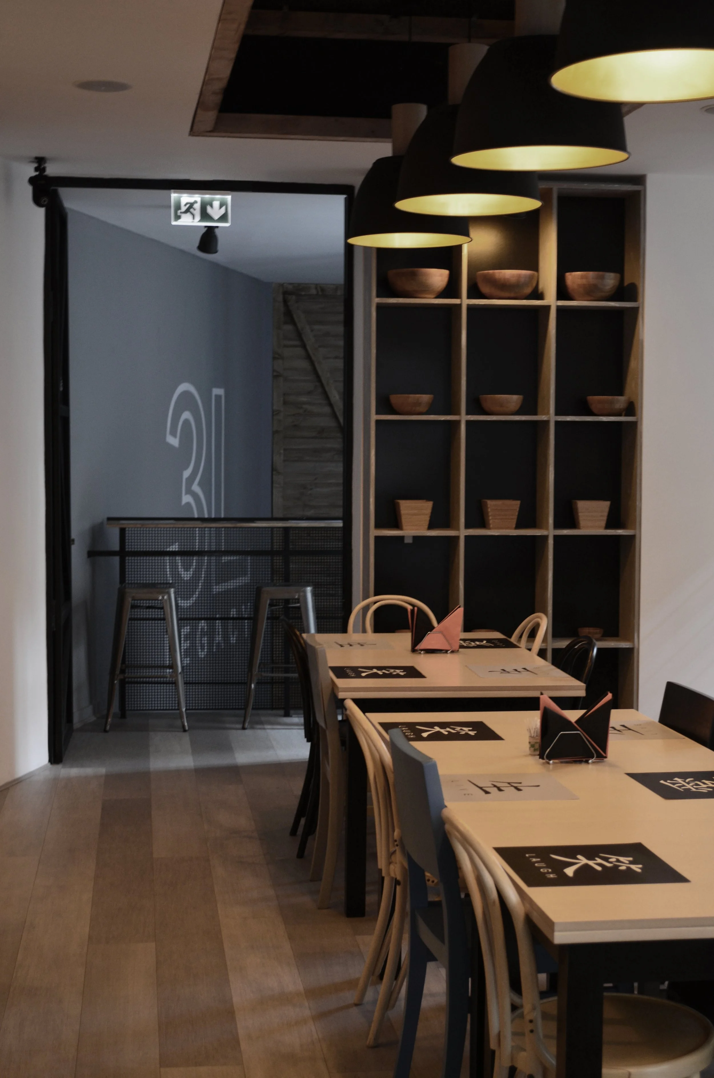 Interior of a modern restaurant with wooden tables, varied chairs, and a shelving unit with wooden bowls and containers, illuminated by hanging pendant lights.