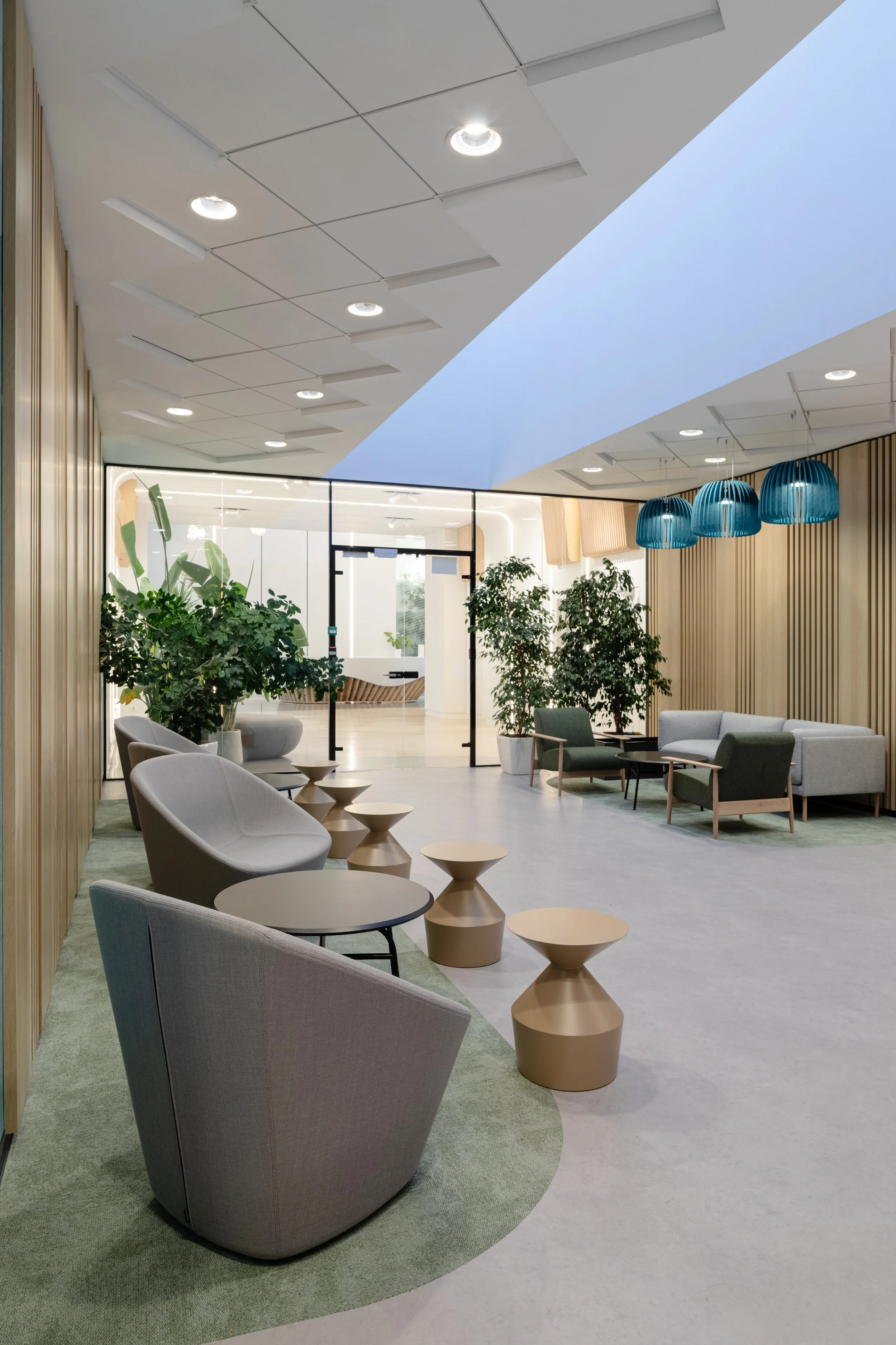 Modern office lobby with gray chairs, beige side tables, green plants, wooden wall panels, blue pendant lights, glass doors.