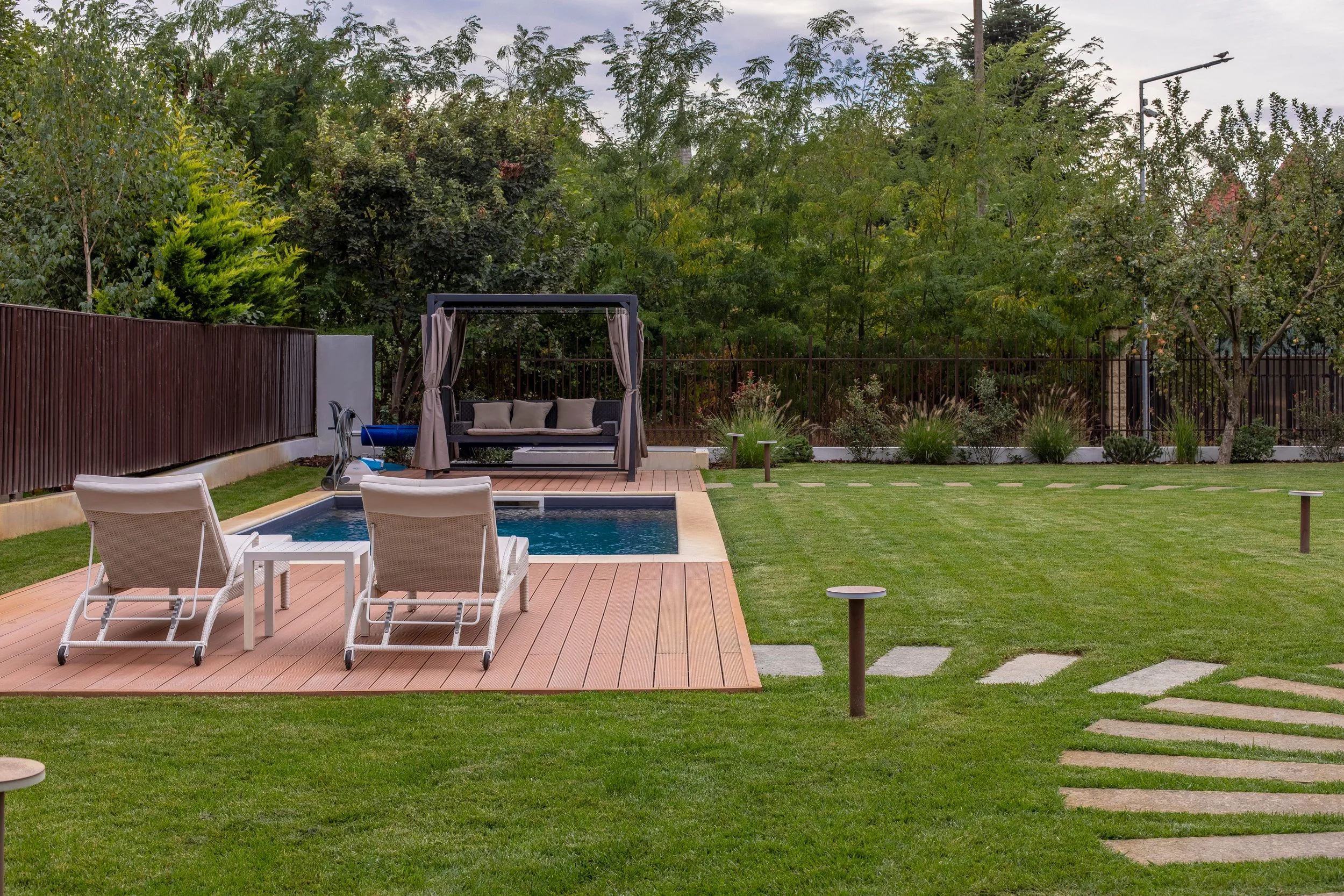 Backyard with a small pool, two lounge chairs, a shaded outdoor swing with cushions, and a grassy lawn with stepping stones, surrounded by trees and a fence.