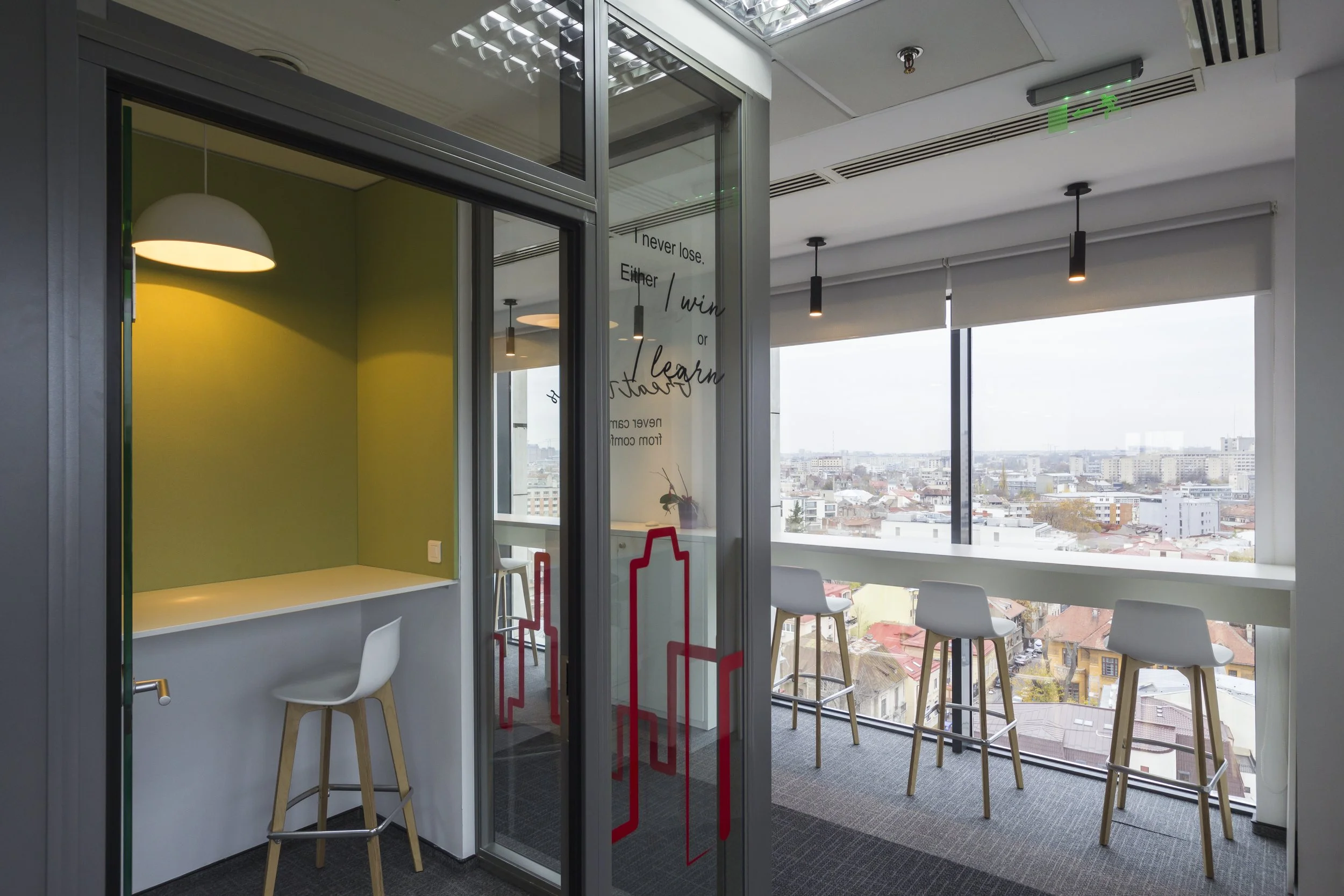 Modern office space with a small seating area, white bar stools by a high window overlooking city buildings, and a cozy corner with a white chair and pendant light.