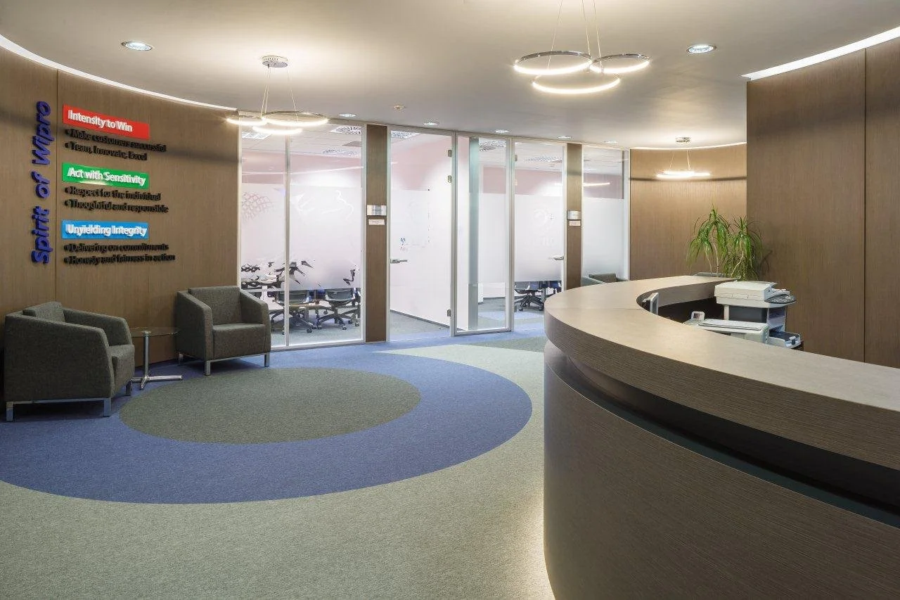 Modern office reception area with curved wooden desk, two chairs, glass conference rooms, and a plant on the right.