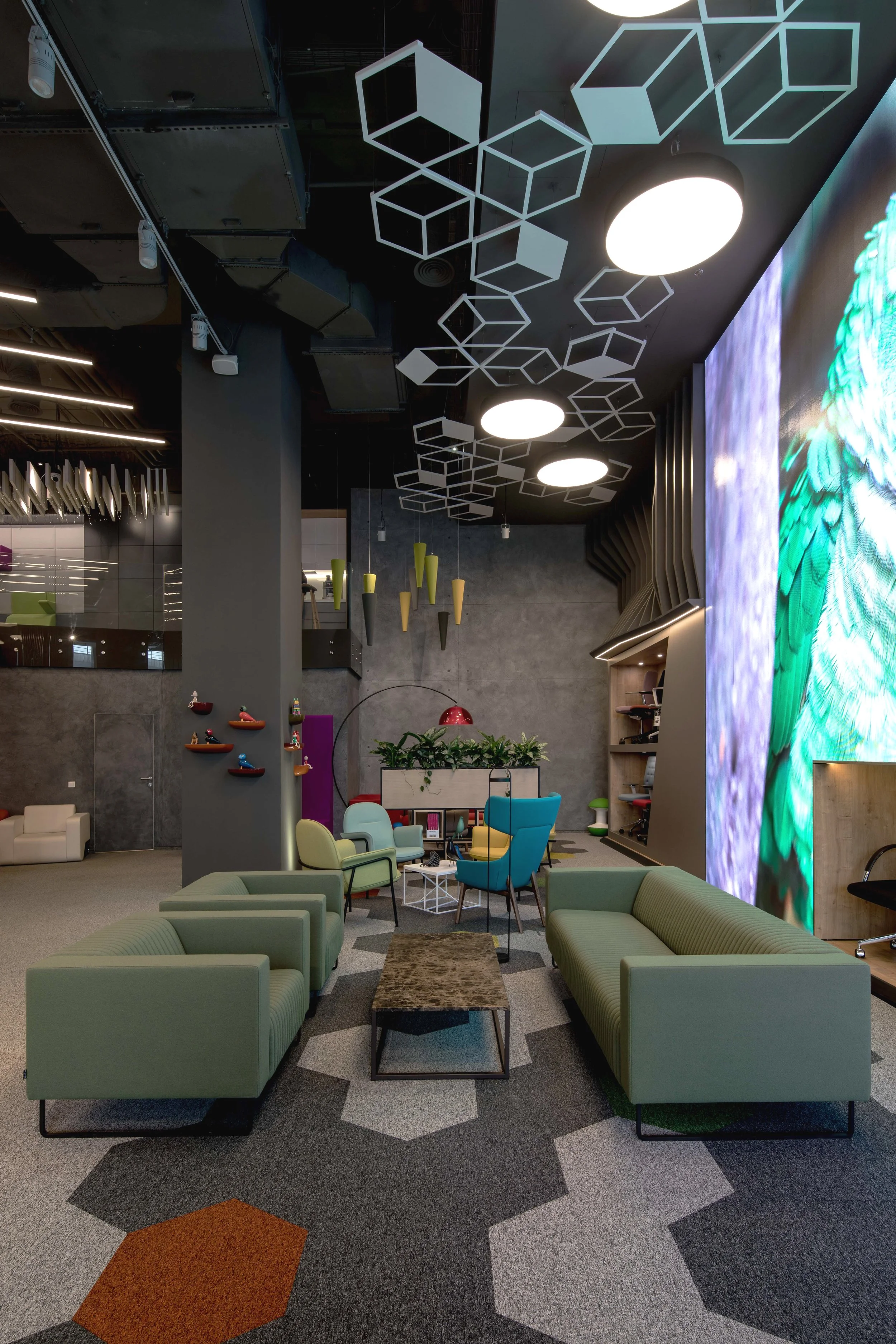Modern lobby with green sofas, colorful chairs, a large digital screen displaying a bird, and geometric ceiling decorations.