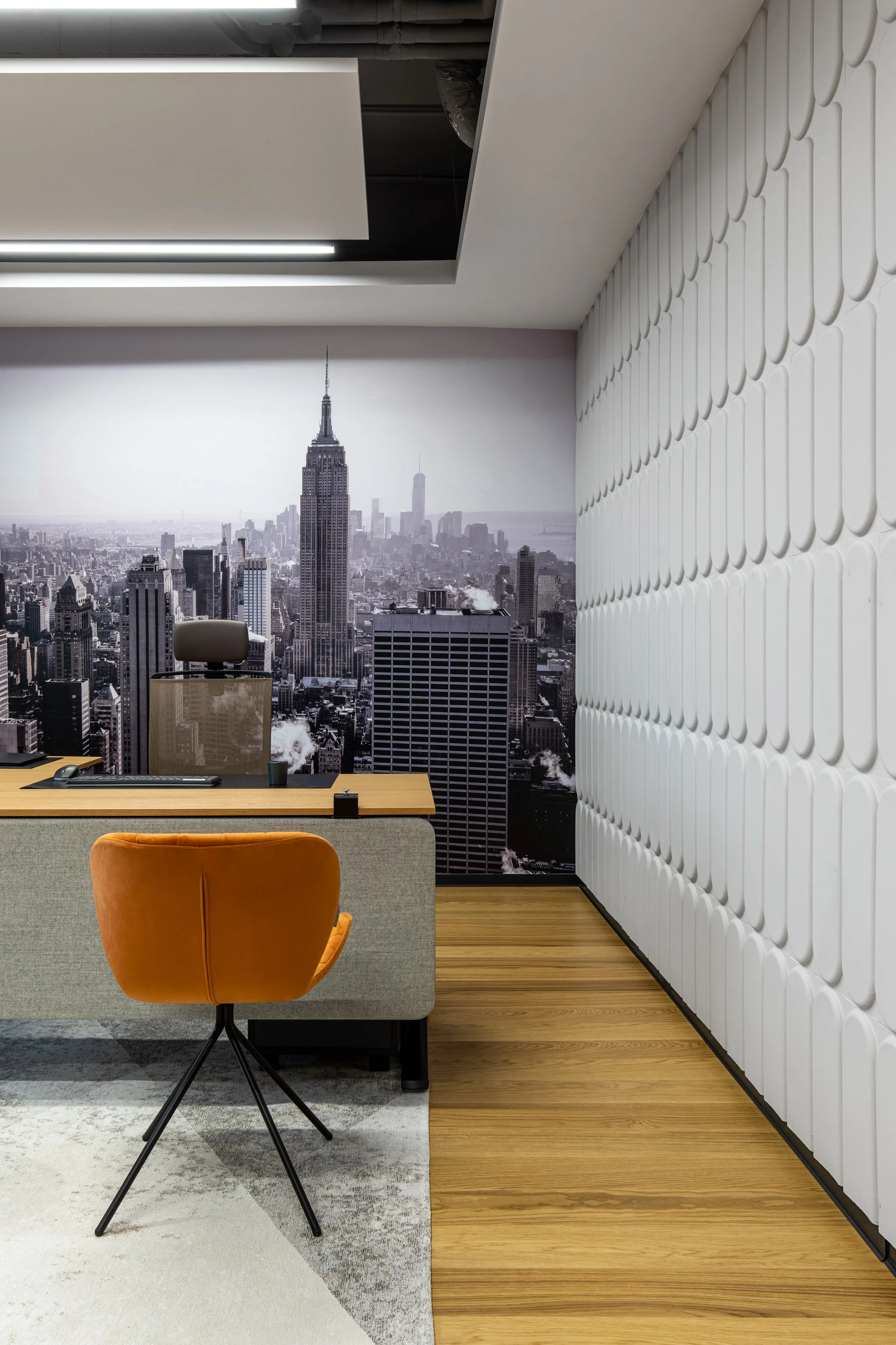Modern office with a desk and an orange swivel chair, a cityscape mural of New York City on the wall, light wood flooring, and white patterned acoustic wall panels.