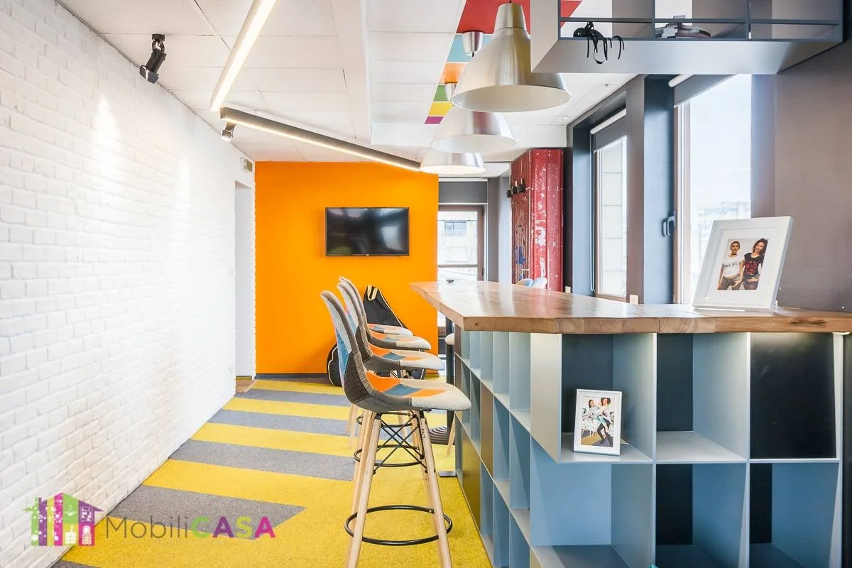 Interior of a modern kitchen or cafe with a colorful and contemporary design. Features a white brick wall, a bright orange accent wall with a mounted TV, and a wooden counter with bar stools. The floor has yellow and gray striped carpet, and there are large windows allowing natural light. Decor includes framed photographs and hanging pendant lights.