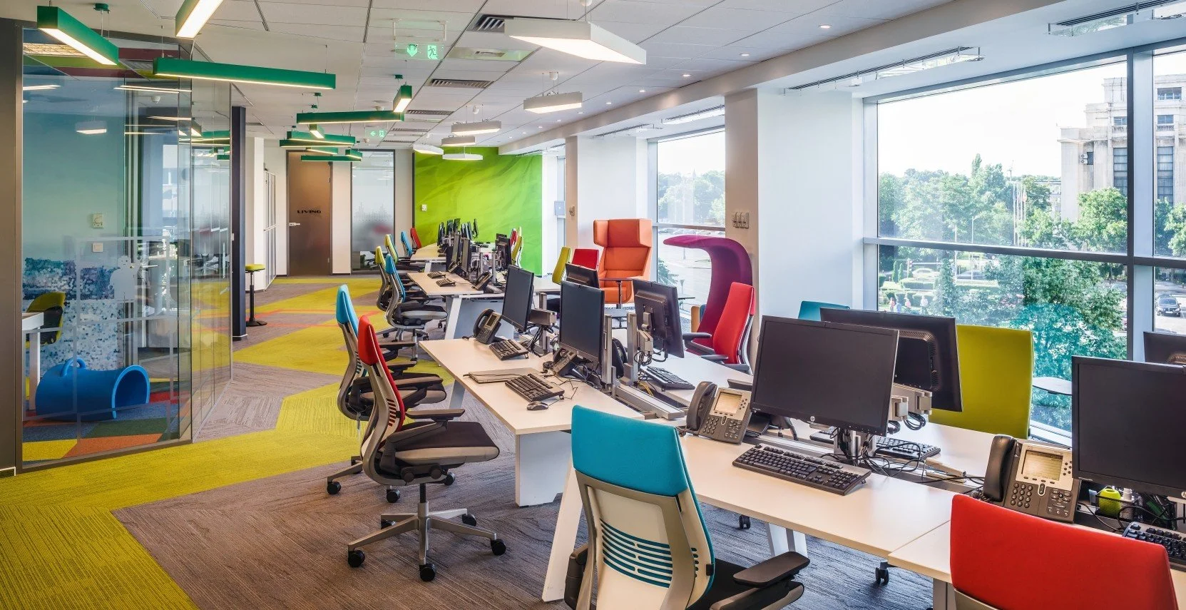 Modern office workspace with multiple desks equipped with monitors, office phones, and chairs in various colors. Large windows provide natural light and a view of trees and buildings outside. Brightly colored wall and ceiling accents create a lively atmosphere.