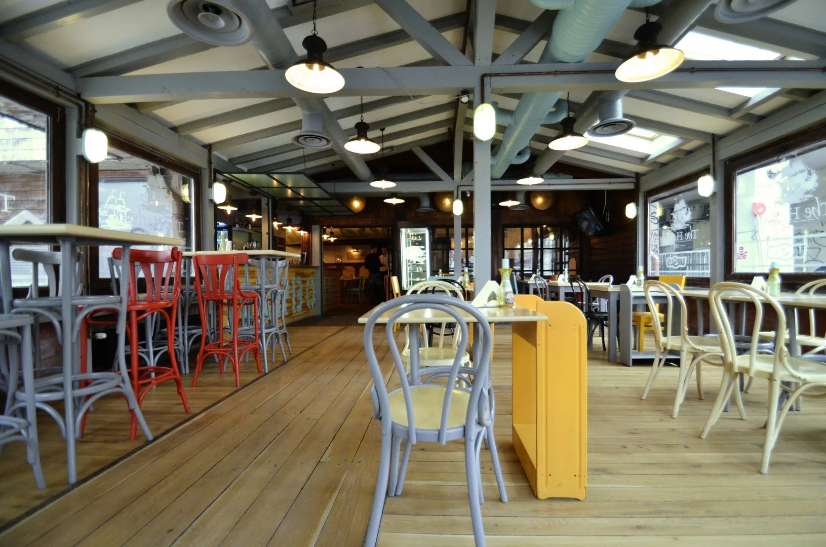 Empty restaurant with wooden floors, colorful chairs, tables set with bottles, large windows, and overhead lights.