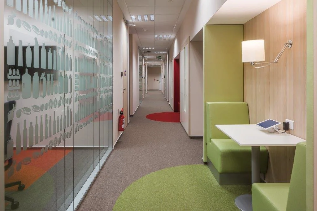 Modern office corridor with colored carpeting, glass walls with frosted designs, and a small seating area with green upholstered booths and a white table, illuminated by a wall-mounted lamp.