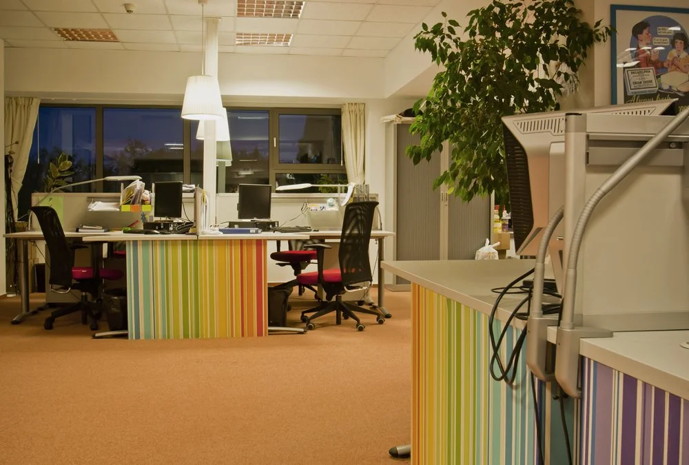 Office space with multiple desks, computers, chairs, a large window, potted plants, and a colorful striped panel on the desks.