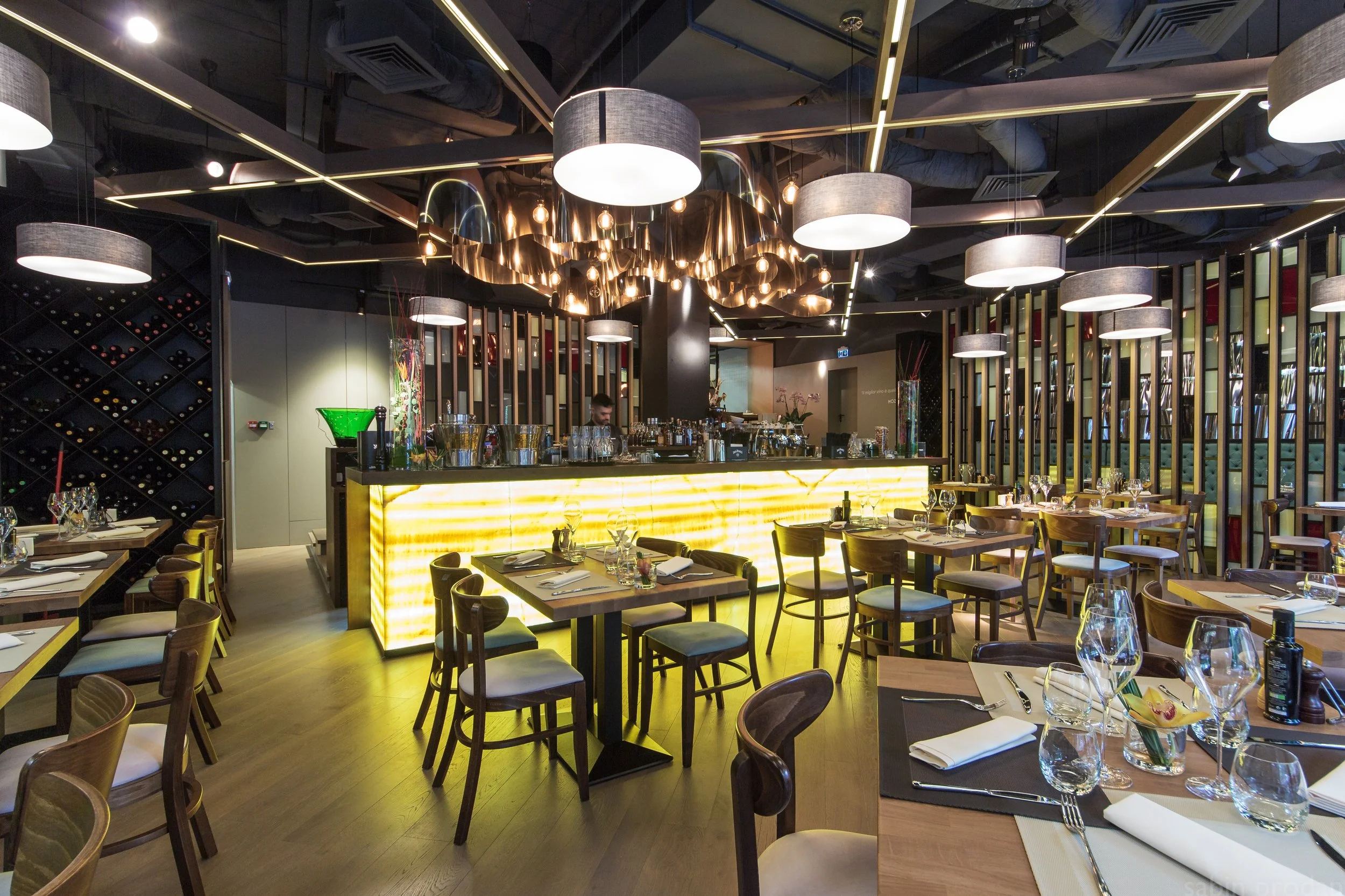 Elegant restaurant interior with modern lighting, a lit yellow bar, wooden tables set with glassware and utensils, and a barista behind the counter.