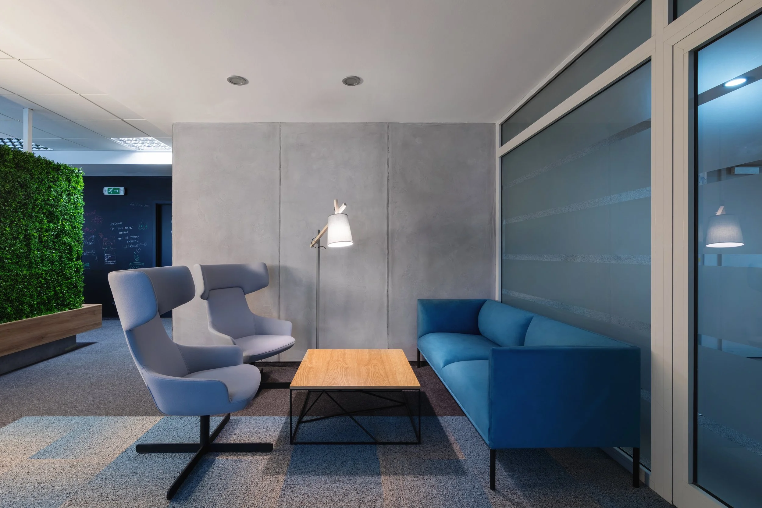 Modern office lounge with blue sofa, two gray armchairs, a wooden coffee table, a floor lamp, and a concrete wall. There's a green wall to the left and office space with glass walls to the right.
