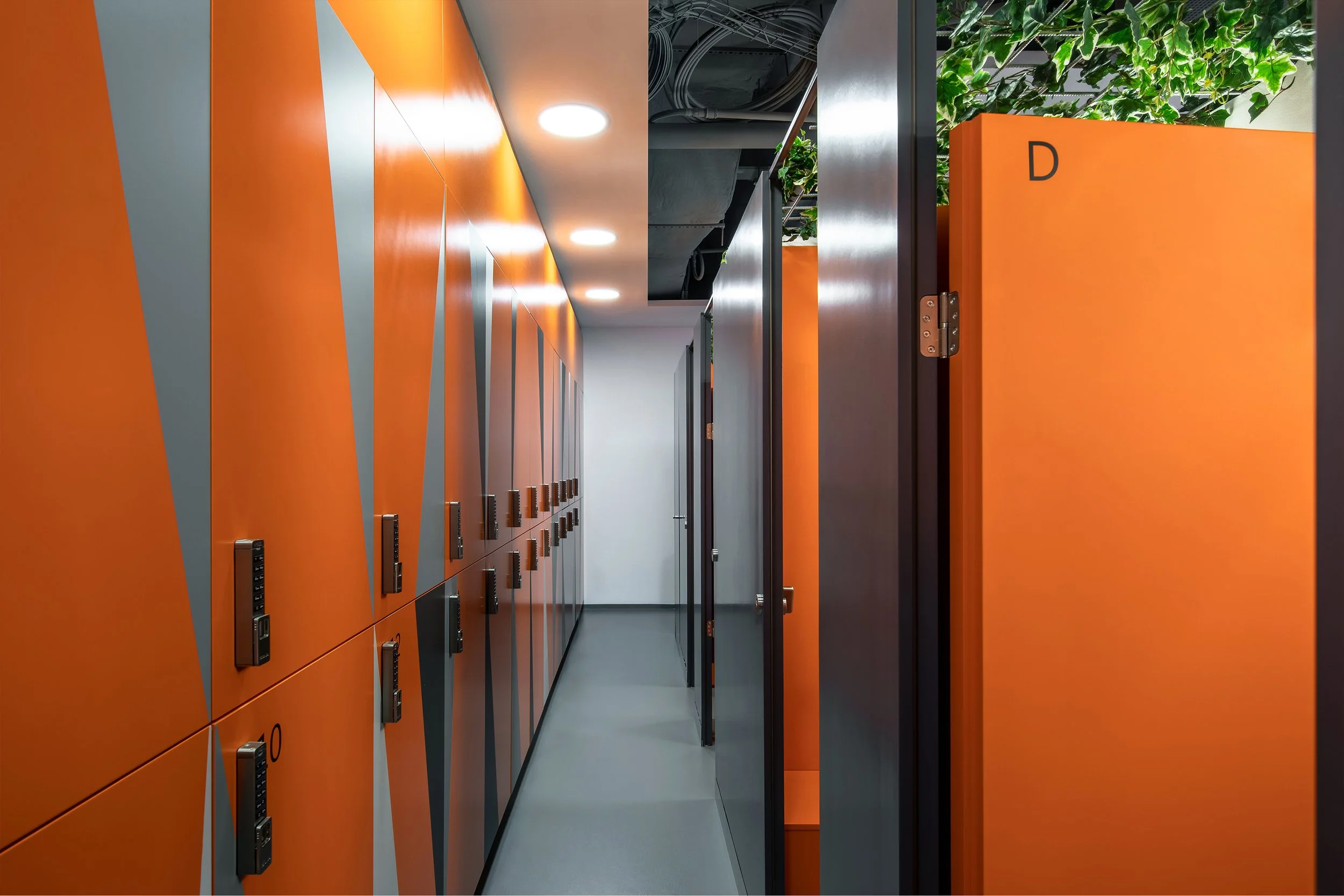 A hallway with bright orange lockers on the left side and orange storage cabinets on the right side, with plants and ceiling pipes visible at the top.
