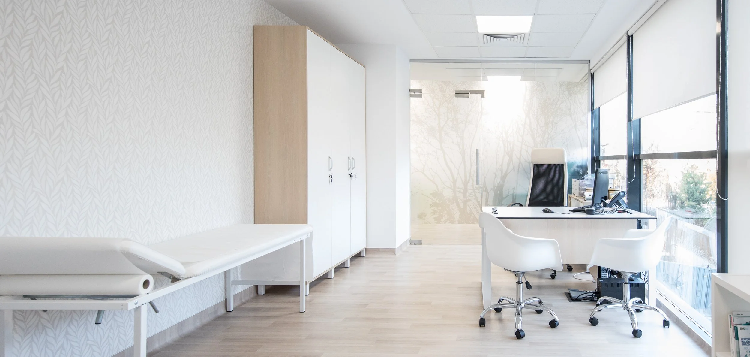 Empty medical consultation room with exam table, cabinets, and a desk with chairs near large windows.