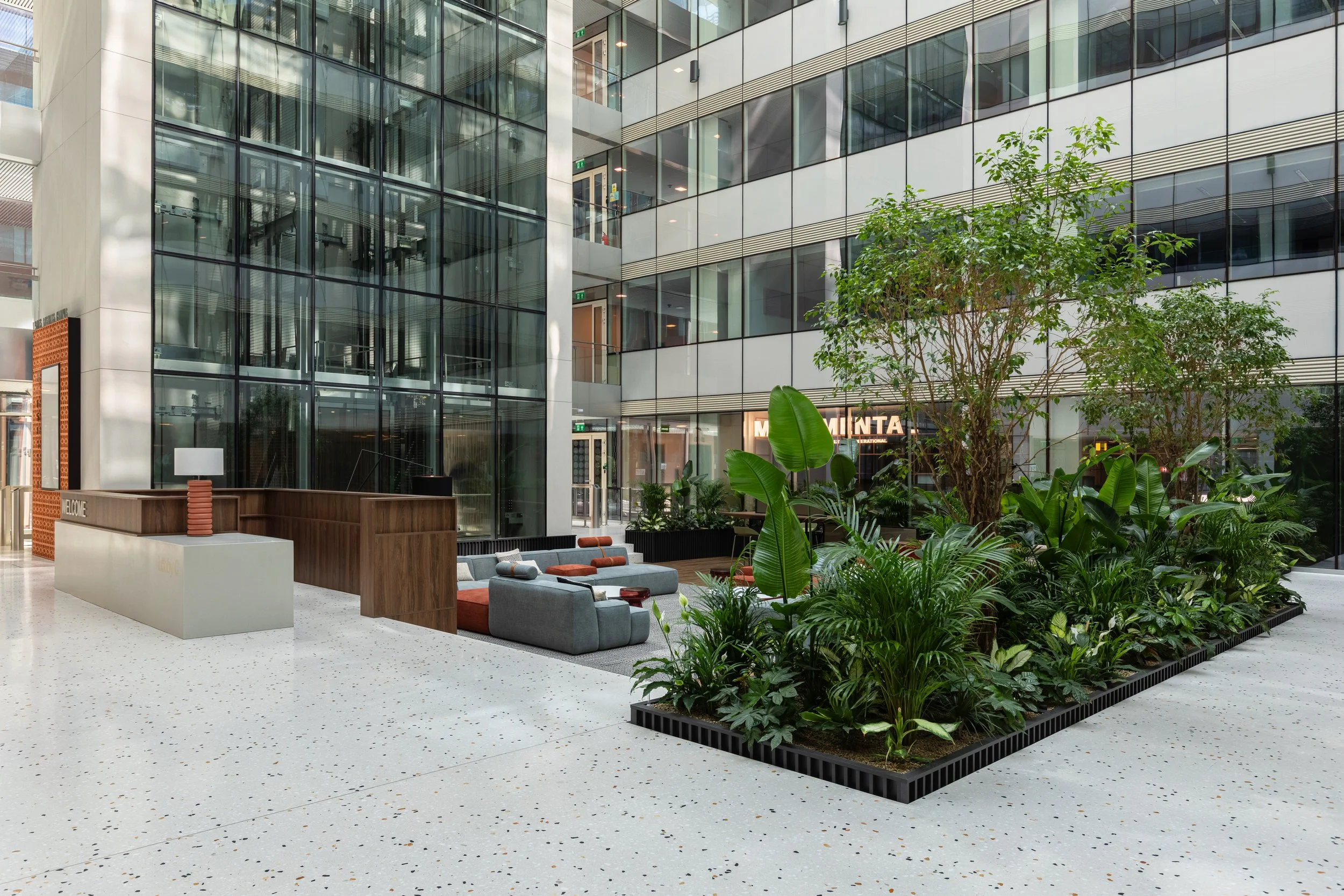 Modern office building lobby with glass walls, indoor plants, and seating area with gray and brown couches.