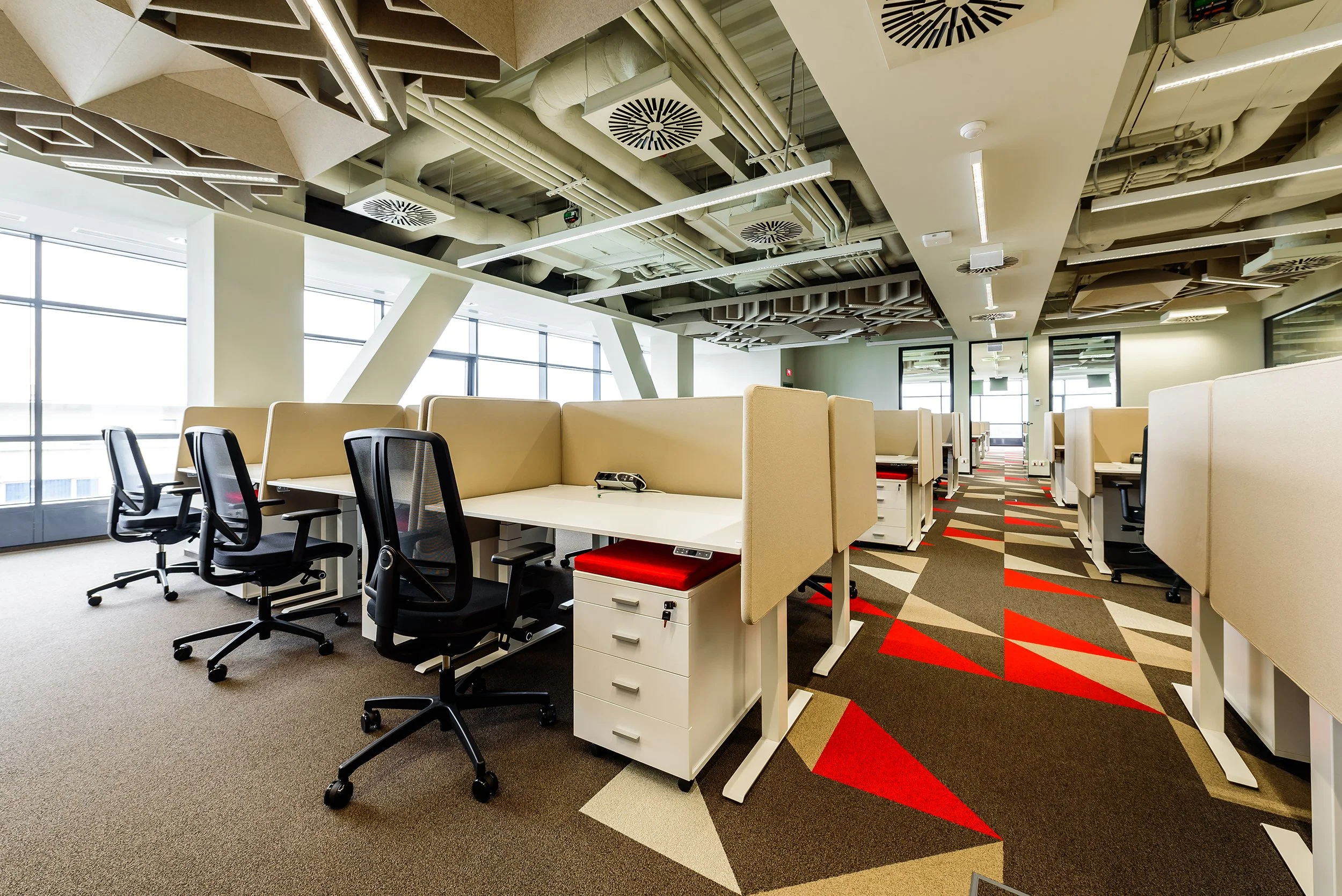Modern open-plan office with workstations separated by beige privacy panels, black office chairs, and a geometric patterned carpet with red, beige, and brown shapes. Large windows in the background allow natural light.