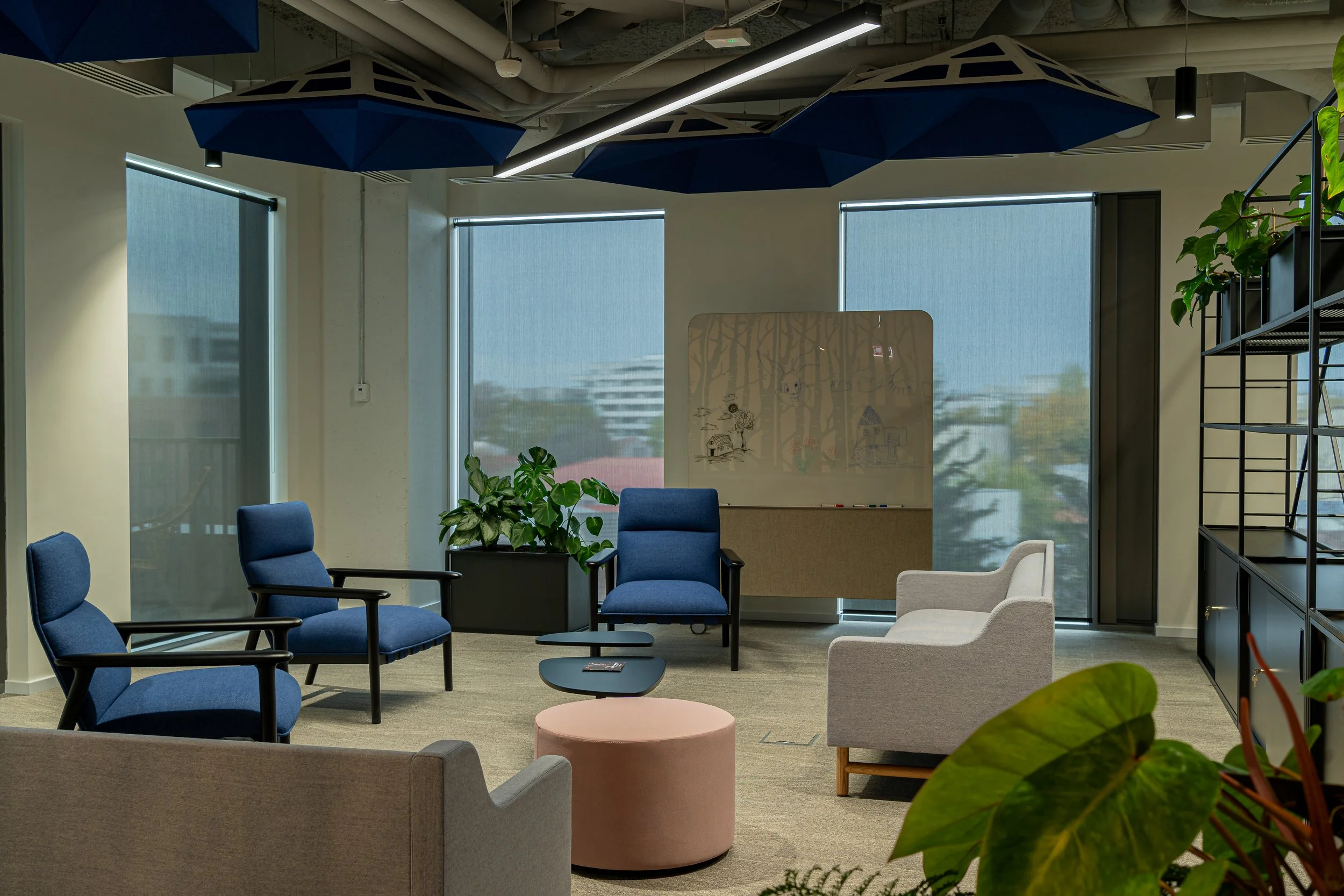 A modern office lounge with blue armchairs, a gray armchair, a round pink ottoman, a small black coffee table, and potted plants.