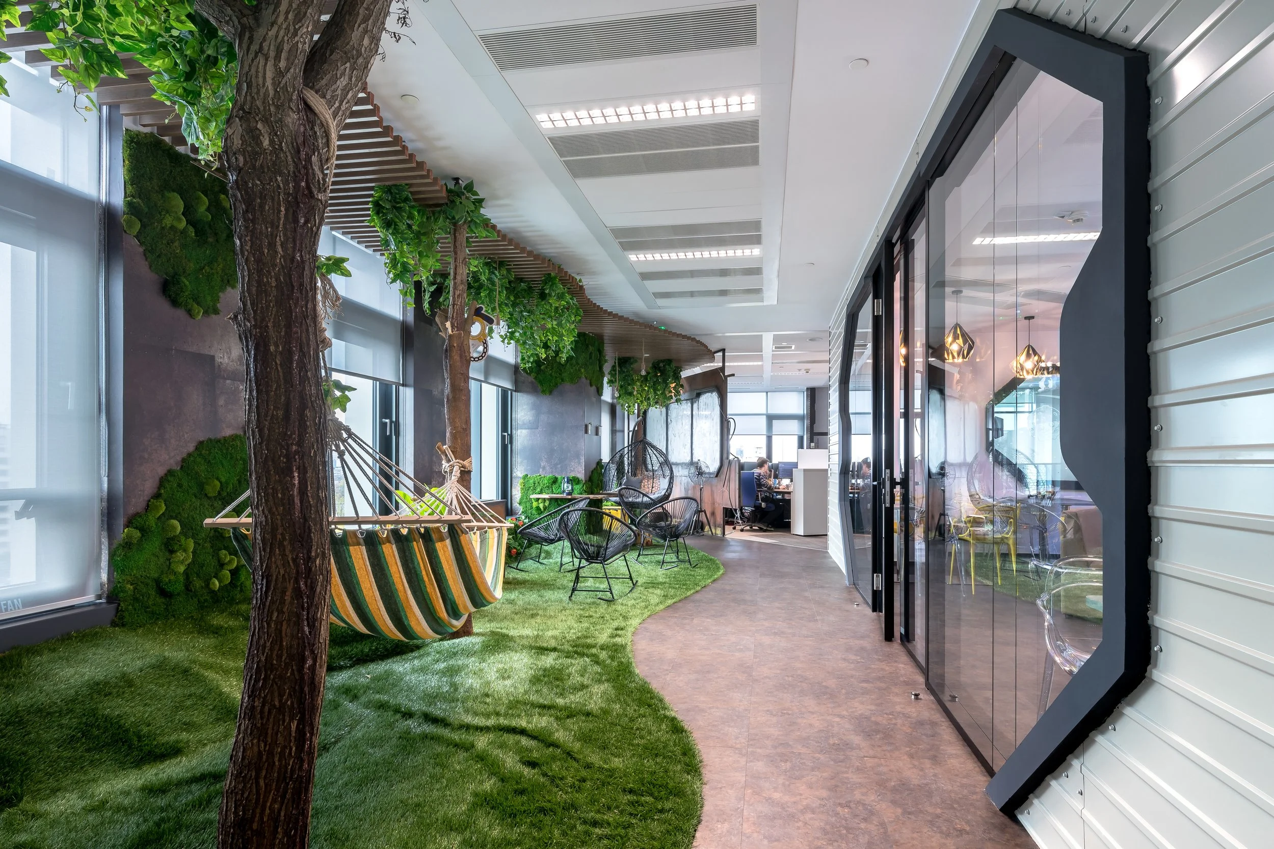 Indoor office space with a green, nature-inspired lounge area featuring trees, hanging plants, a hammock swing, and chairs on artificial grass, contrasted with a modern conference room with glass walls on the right.