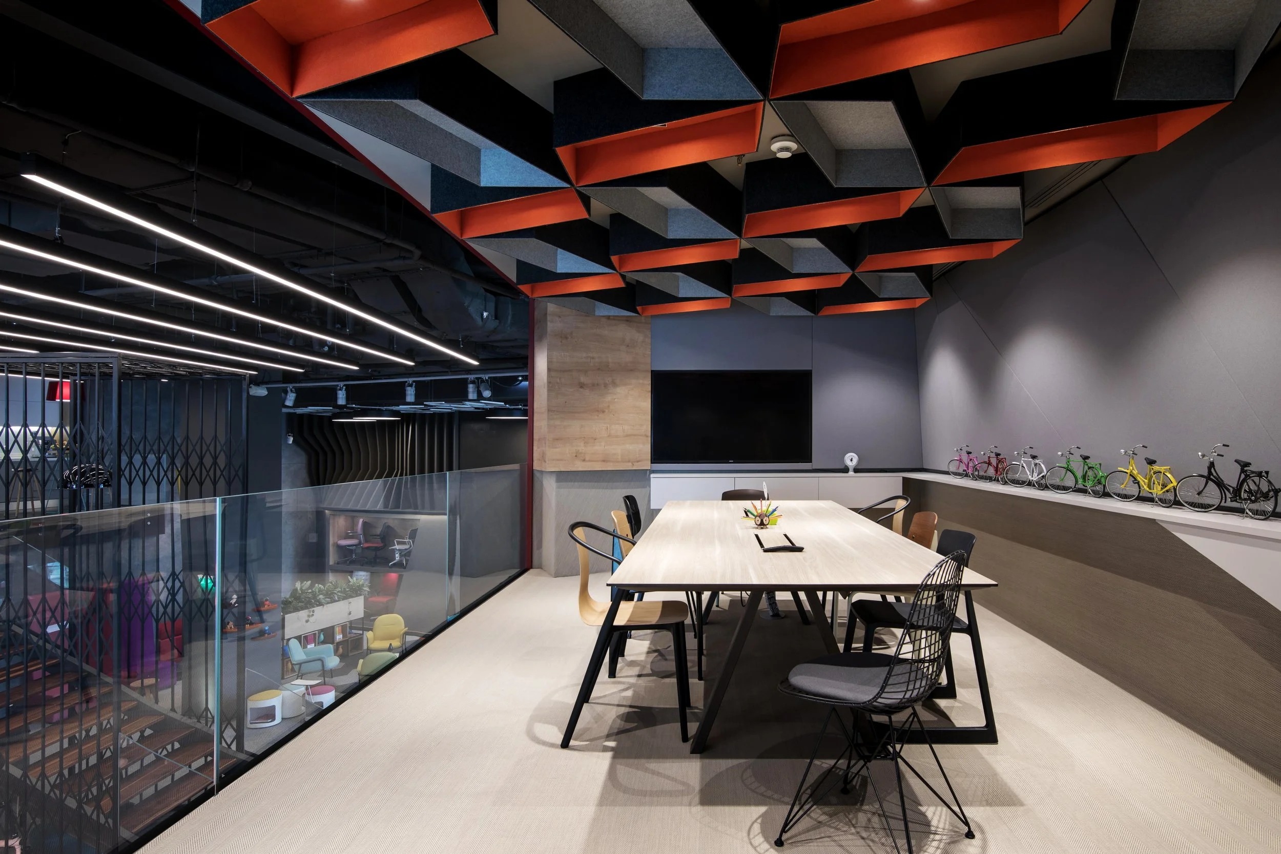 Modern office meeting room with a long light-colored wooden table, chairs, a wall-mounted TV, and small colorful bicycles displayed on a shelf, with a glass partition overlooking a vibrant office space below.