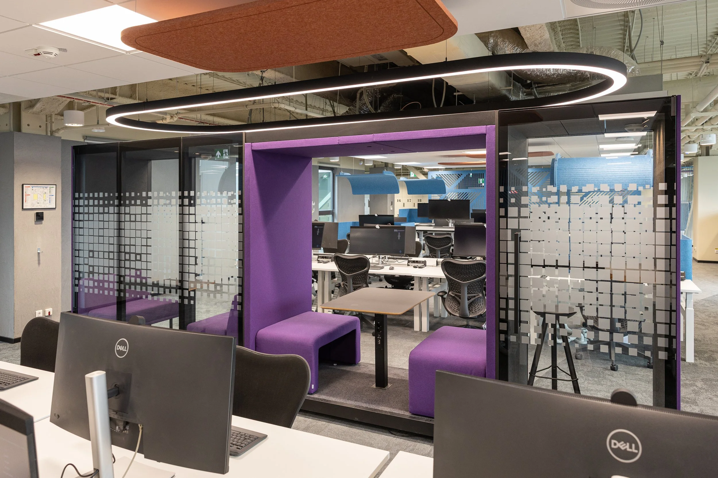 Modern office space with a purple enclosed meeting nook, glass walls with pixelated pattern, black ergonomic chairs, computer desks, and ceiling lighting