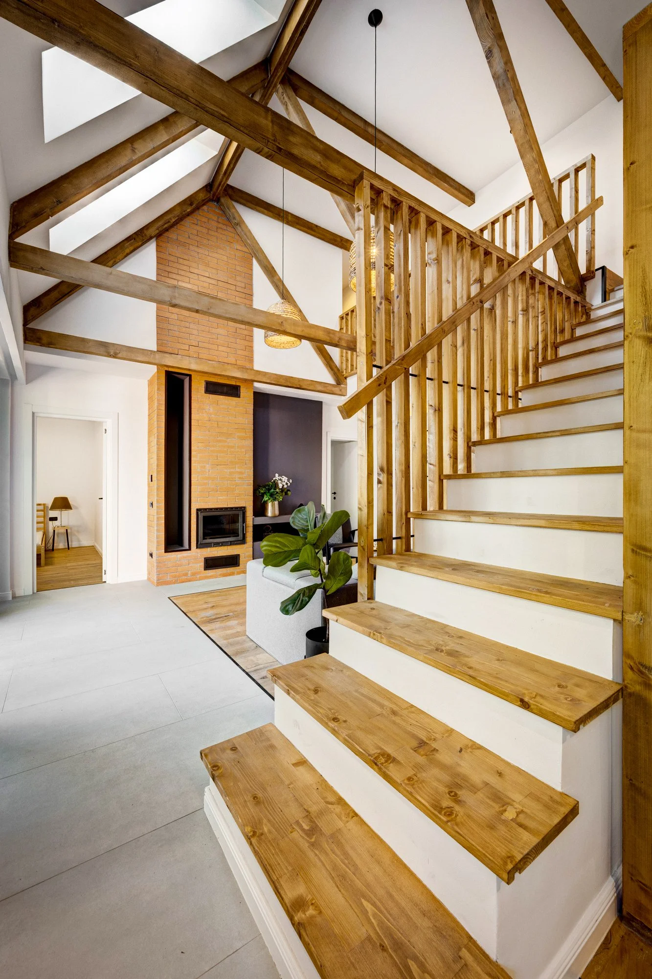 Interior view of a modern house featuring a wooden staircase and exposed wooden beams, with a brick fireplace and a sitting area.