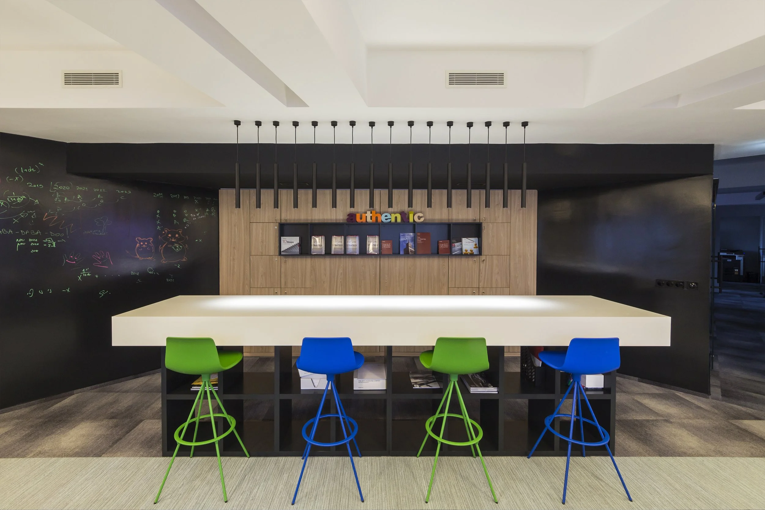 Modern office lounge with a white bar-height table, four colorful chairs (green and blue), black shelving underneath, a wooden accent wall with the word 'authentic' in colorful letters, a blackboard wall with chalk drawings of animals and notes, and ceiling lights hanging above.