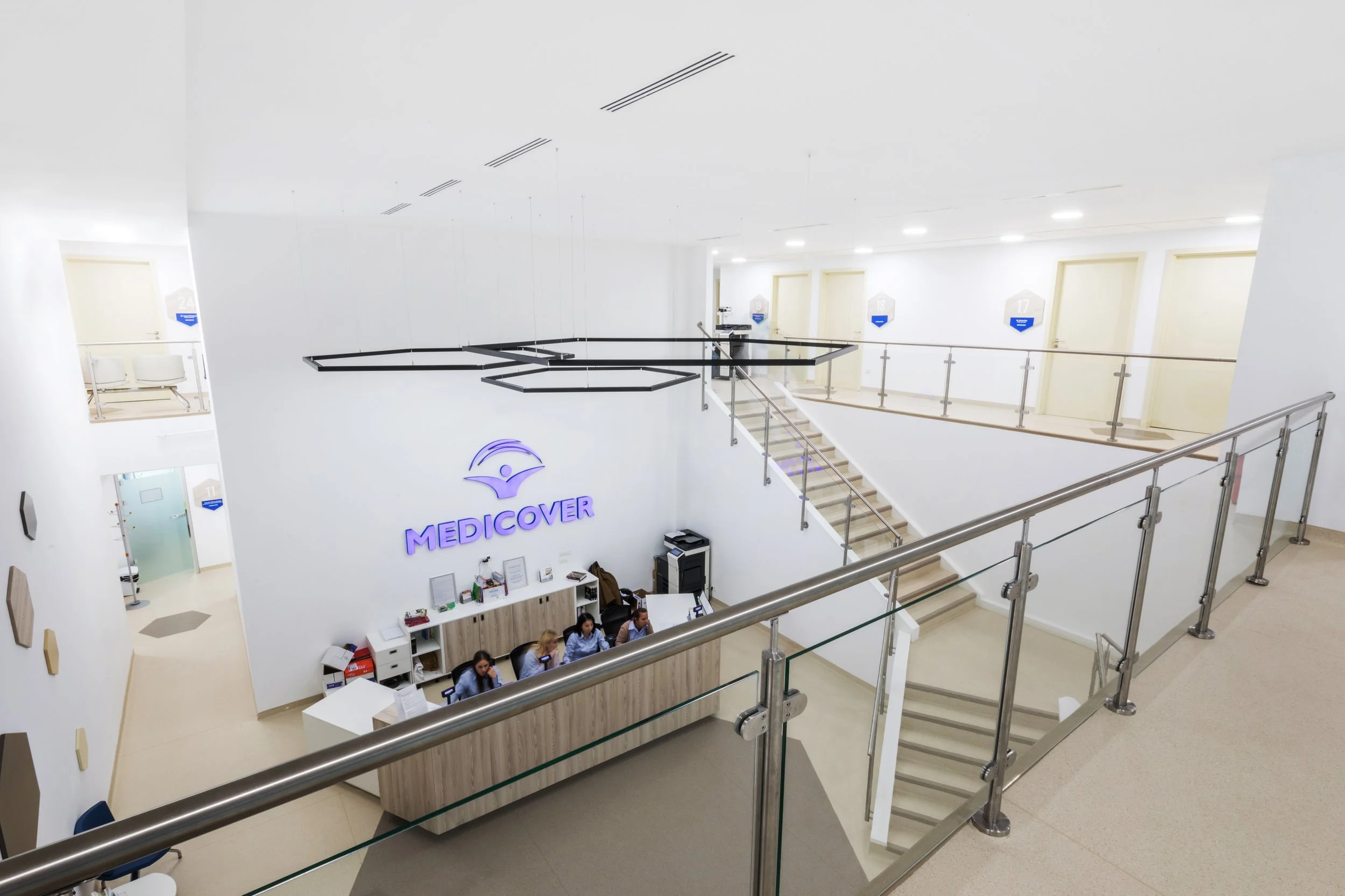 Modern Medical Center reception area with staff seated behind a curved counter, illuminated 'MEDICOVER' signage on the wall, and multiple doors labeled with numbers in the background.