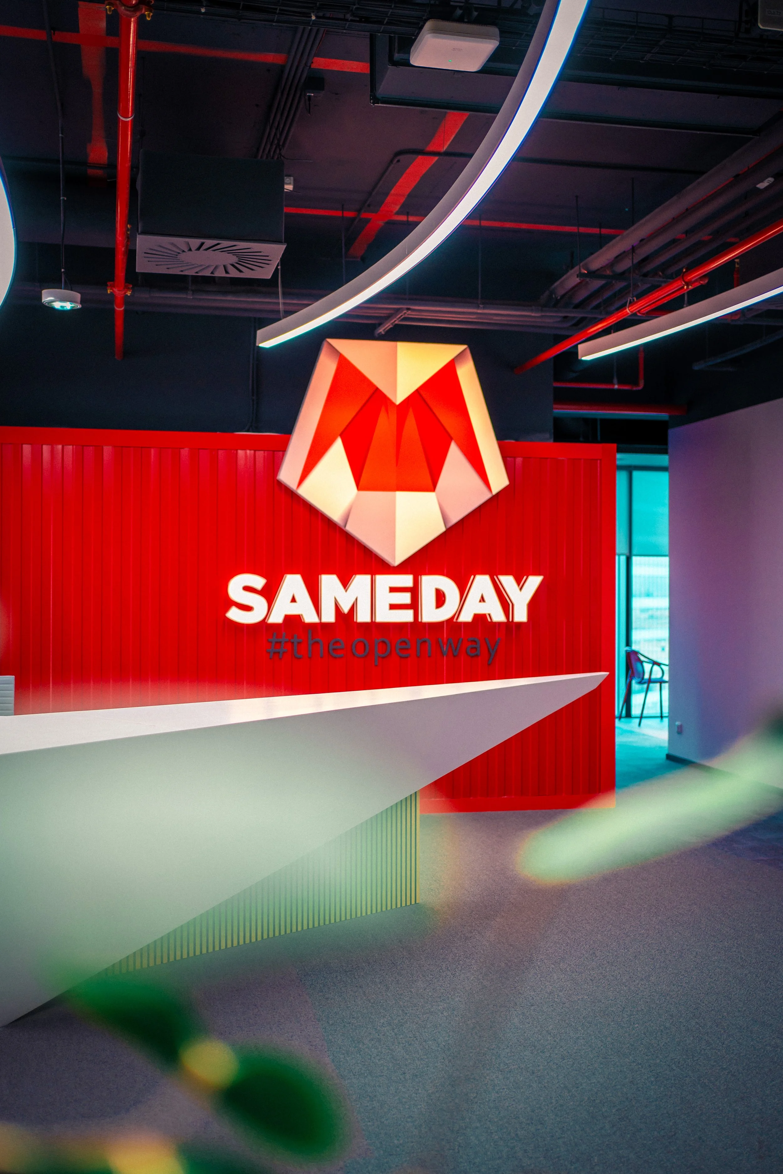 Indoor reception area with a red wall featuring a large geometric fox head logo, illuminated text reading "SAME DAY" and hashtag "#theopenway", modern lighting fixtures, and a white angular counter in the foreground.