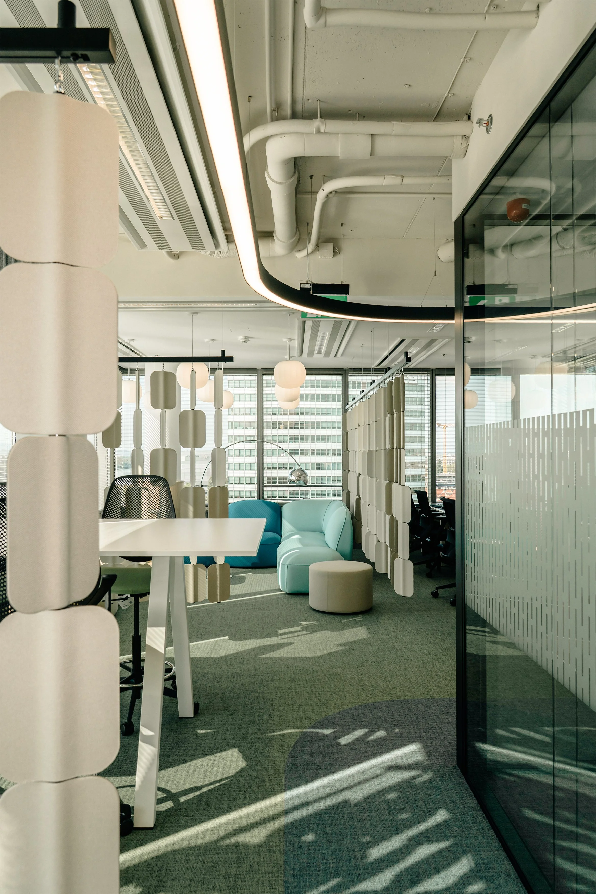 Modern office lounge with light-colored furniture, partition panels, and large windows letting in natural light.