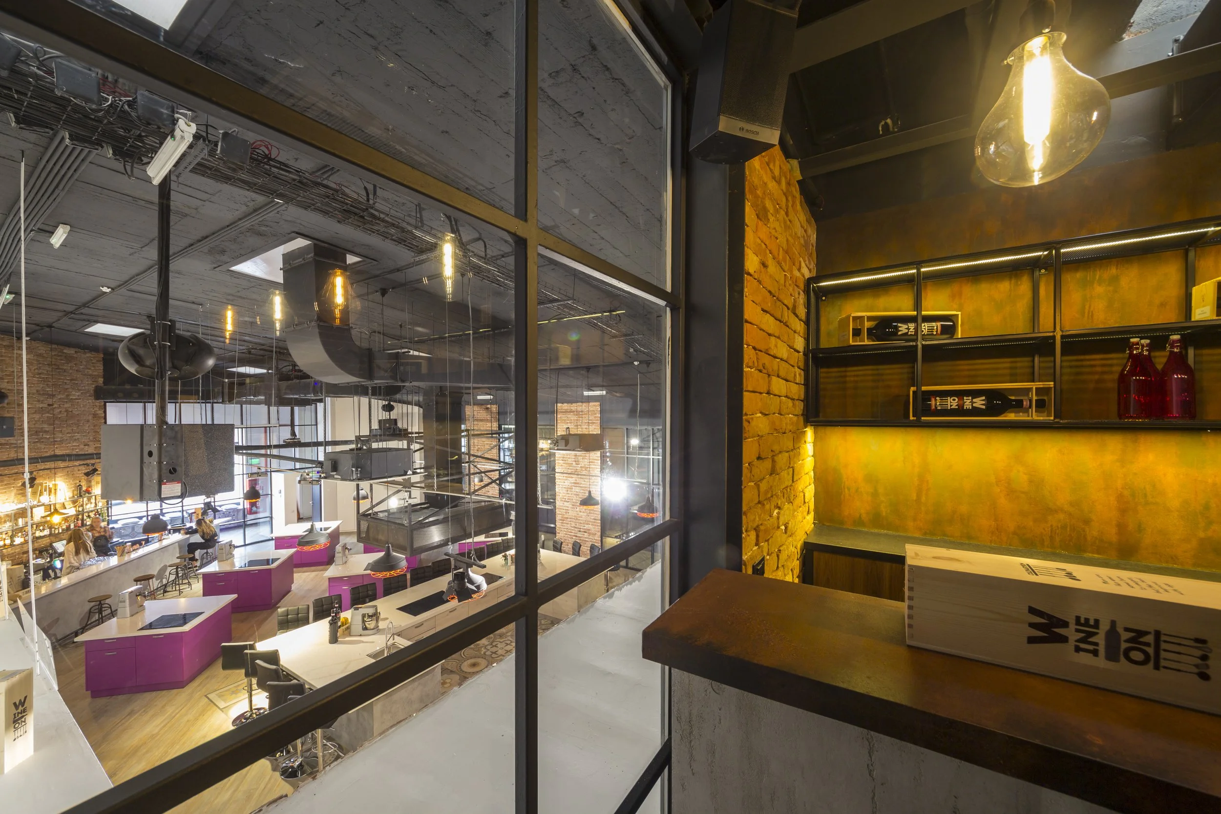 View of a modern, industrial-style café or coworking space with exposed brick walls, large windows, and a seating area with purple booths and stools, seen from a small corner with a wooden countertop and a glass partition.