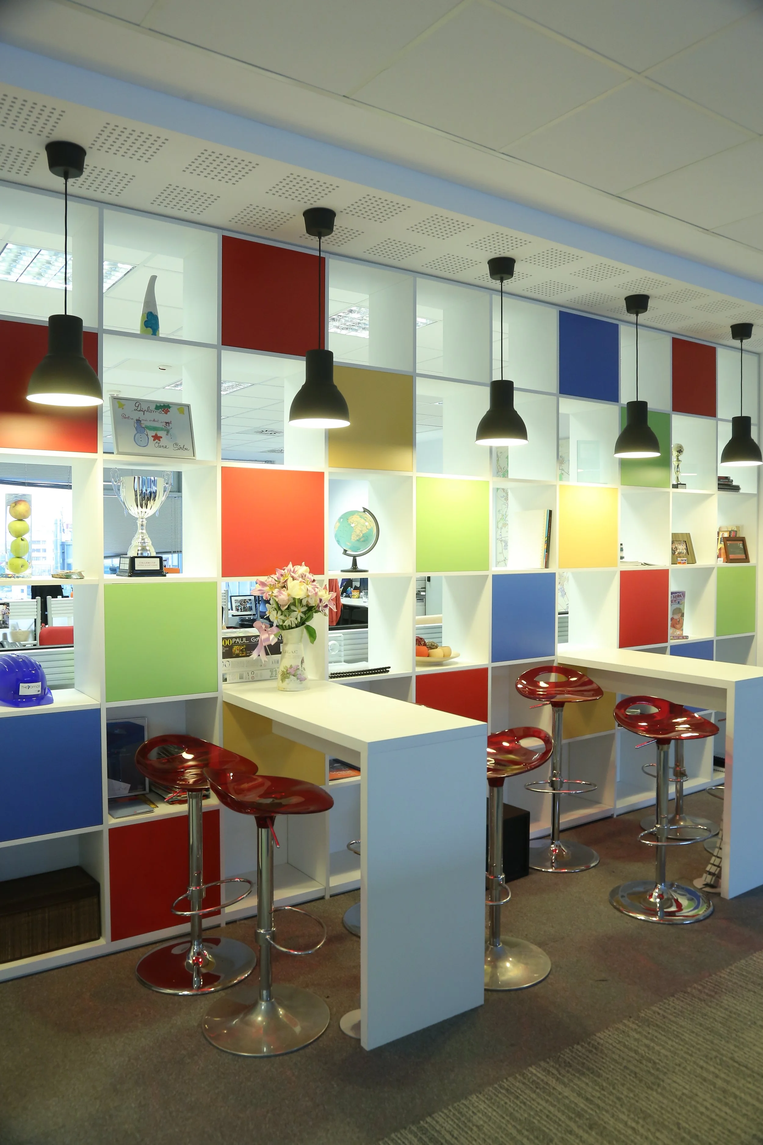 Colorful office lounge with a white shelving unit featuring red, green, blue, and yellow squares, decorated with trophies, a globe, and framed photos. Four red bar stools with chrome bases are positioned at a white high table.