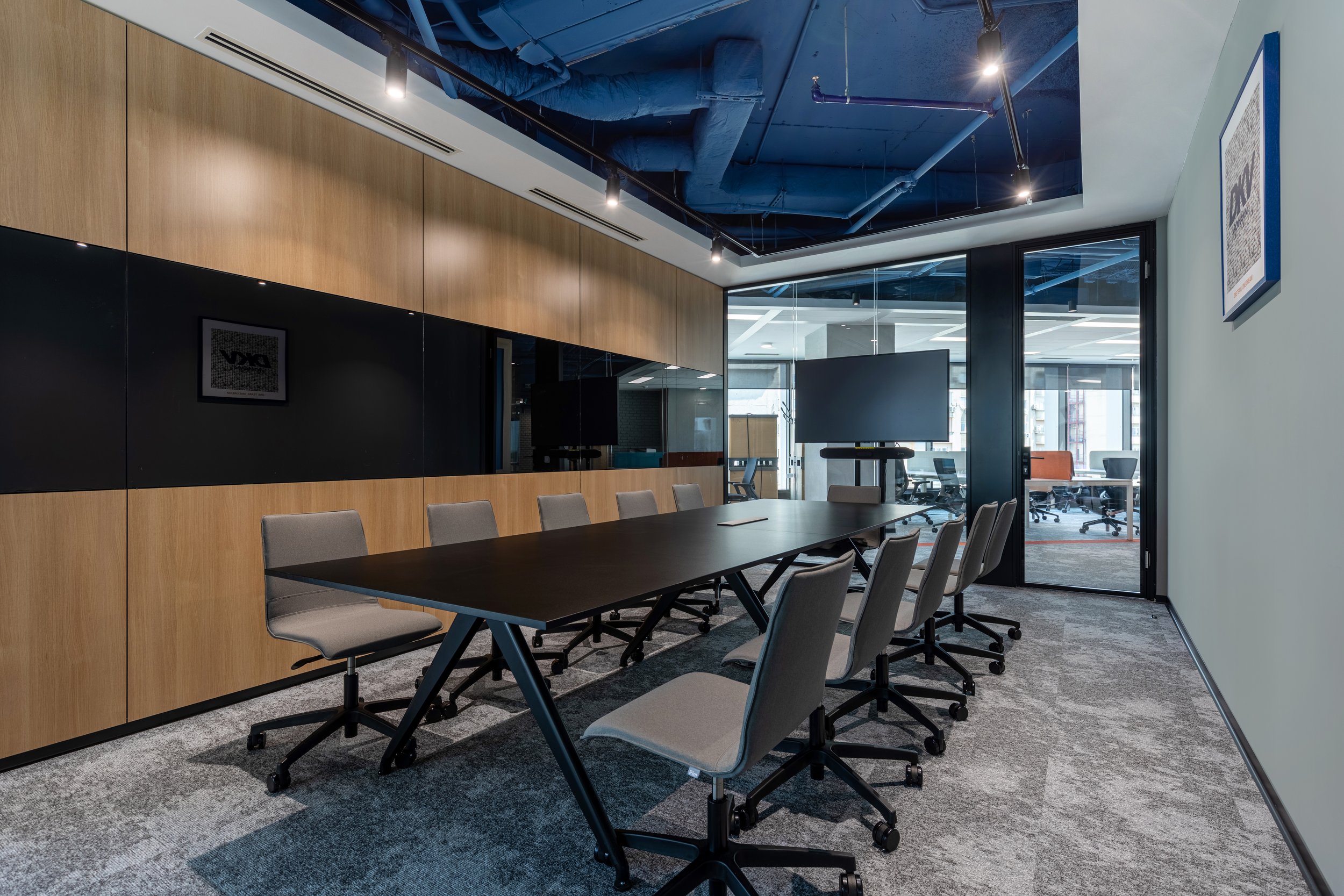 A modern conference room with a long dark table surrounded by gray office chairs. On the far wall, there is a black and wood-paneled wall with a mirror, and a large monitor or screen stands on a mobile cart near the window. The ceiling is painted blue with exposed ductwork and track lighting. The room has large windows with a view into an open office space.